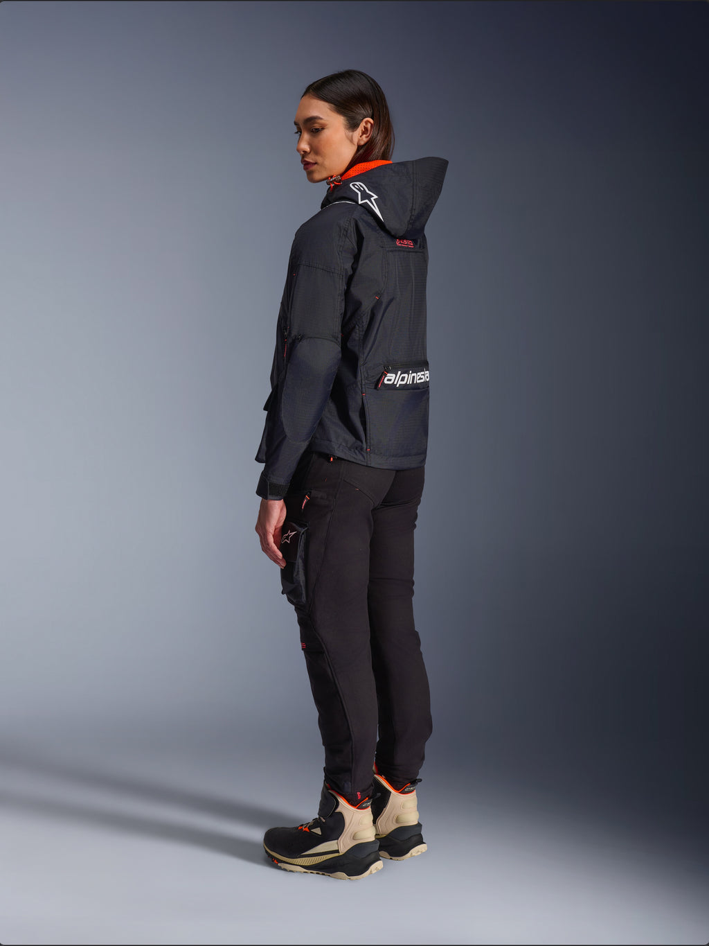 Pantaloni Alpinestars Actea da Donna, nero, moderni pantaloni da moto con vestibilità affusolata e tasca cargo sulla coscia, mostrati da una prospettiva laterale-posteriore su una modella che indossa una giacca scura con cappuccio e stivali da moto, stile urban riding.