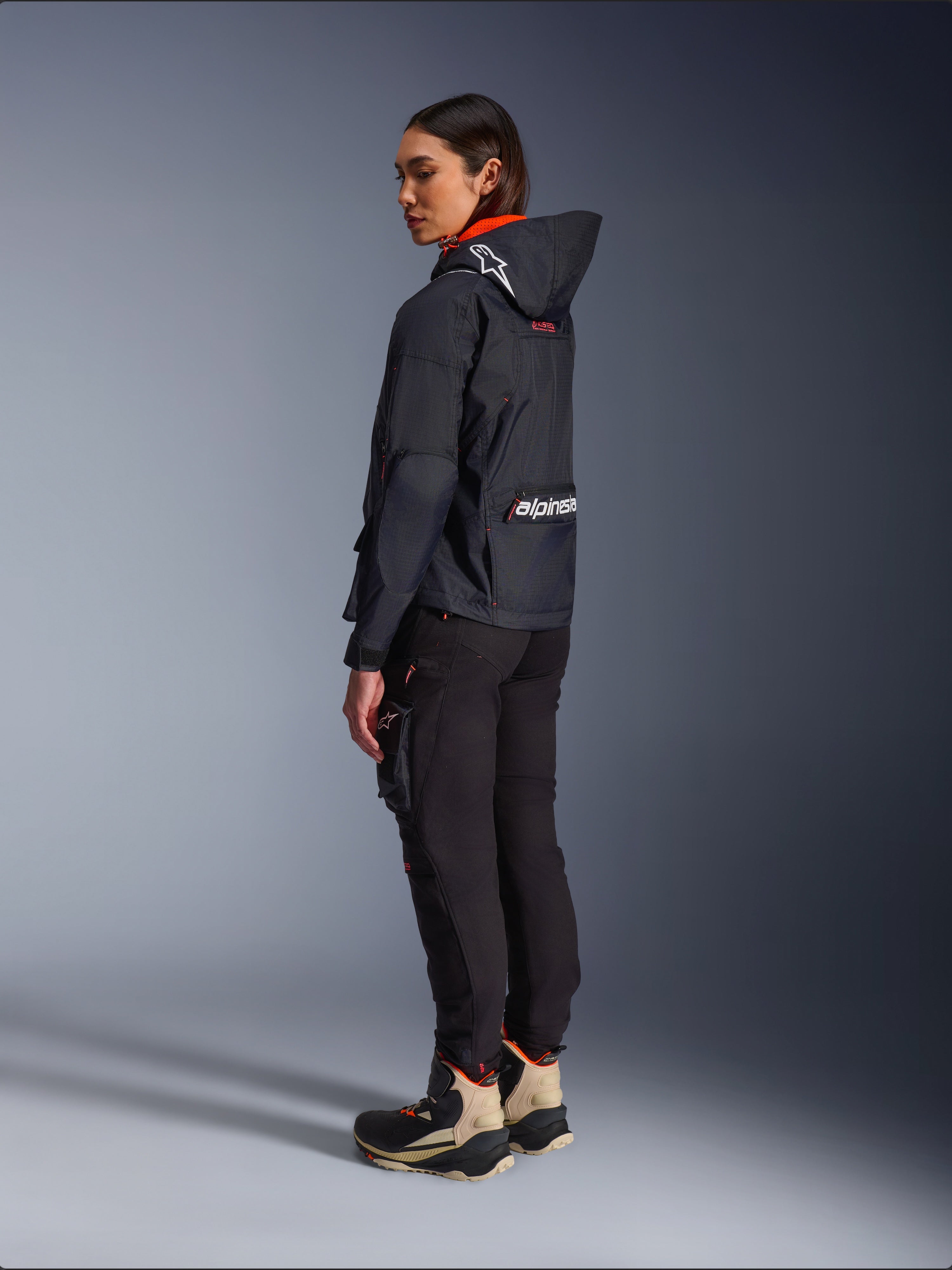 Pantaloni Alpinestars Actea da Donna, nero, moderni pantaloni da moto con vestibilità affusolata e tasca cargo sulla coscia, mostrati da una prospettiva laterale-posteriore su una modella che indossa una giacca scura con cappuccio e stivali da moto, stile urban riding.