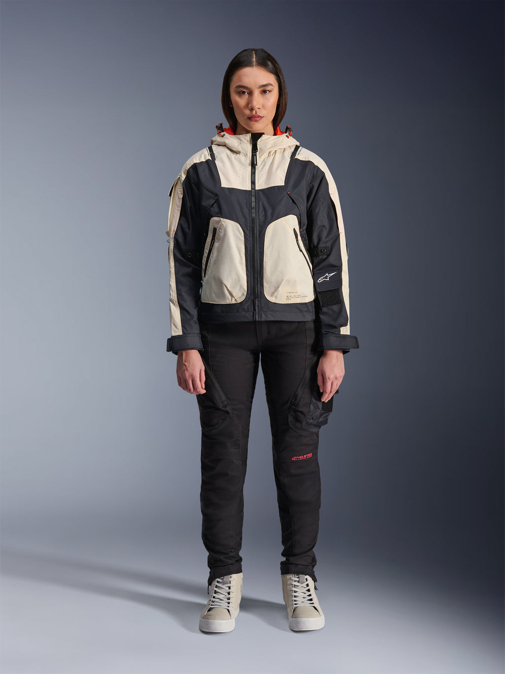 Modella che indossa la giacca Alpinestars Molly Air in Black Almond, con design color-block crema e nero e cappuccio foderato in rosso, abbinata a pantaloni da moto neri e sneakers crema, scatto in studio di abbigliamento urban contemporaneo.