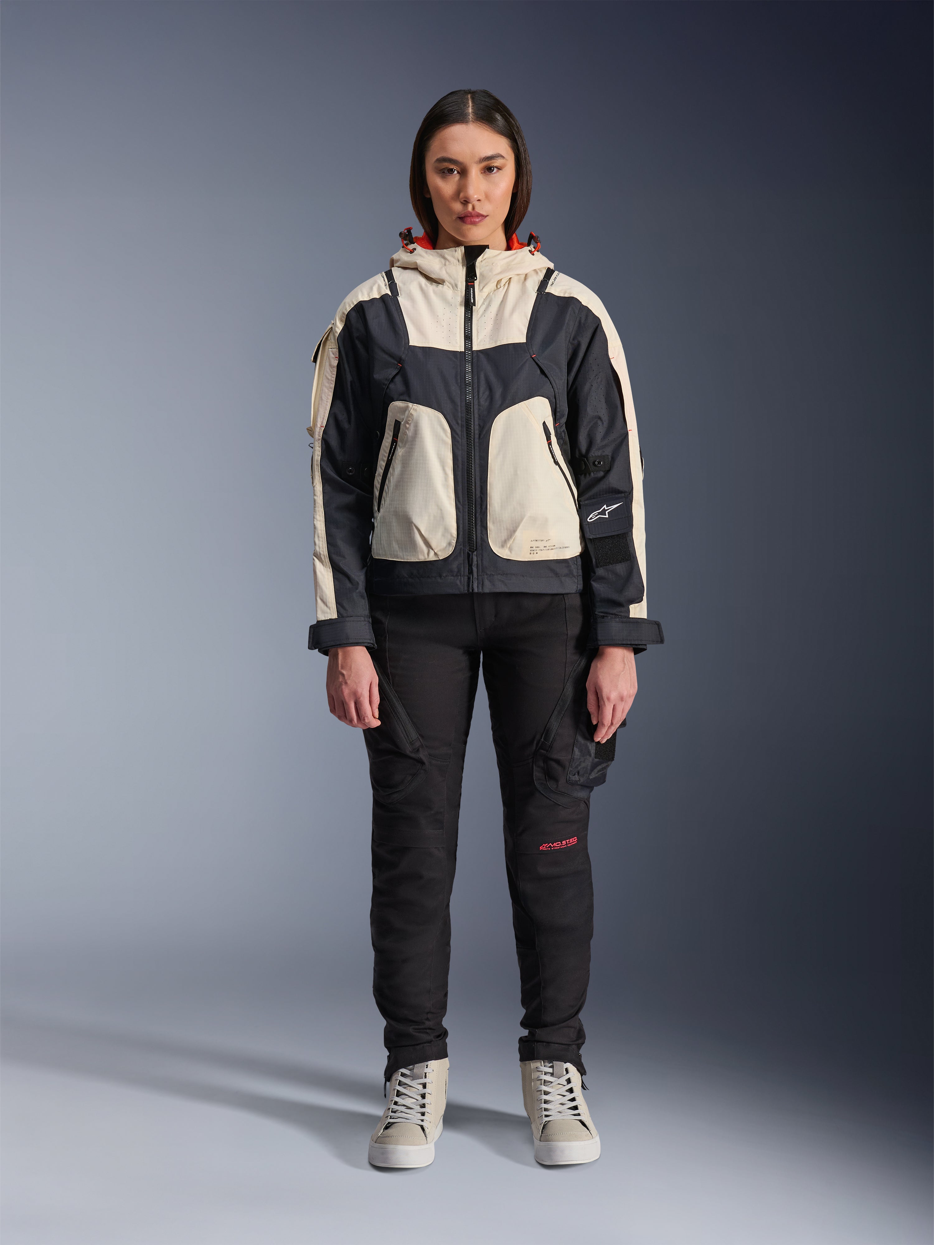 Modella che indossa la giacca Alpinestars Molly Air in Black Almond, con design color-block crema e nero e cappuccio foderato in rosso, abbinata a pantaloni da moto neri e sneakers crema, scatto in studio di abbigliamento urban contemporaneo.