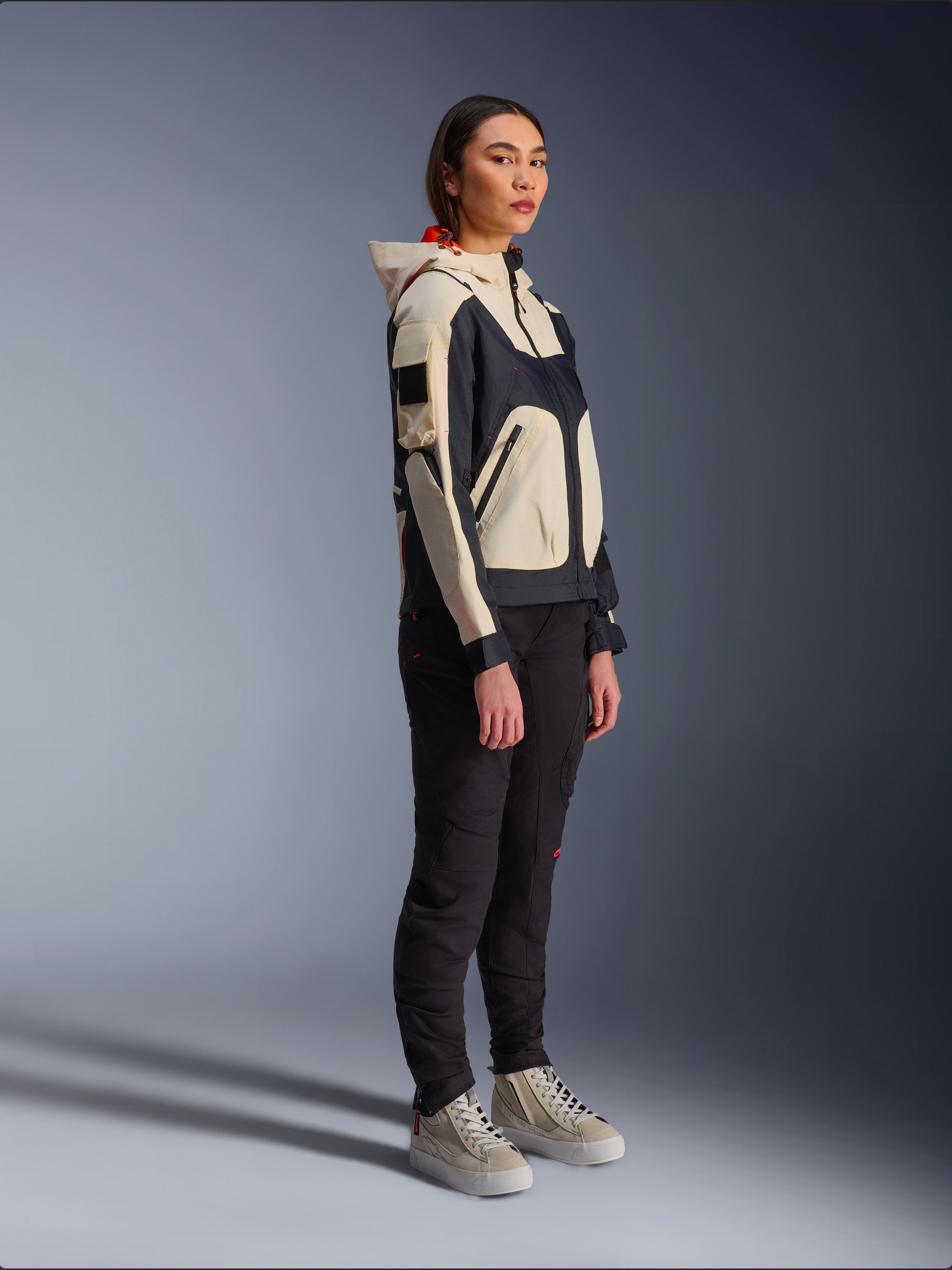 Giacca Alpinestars Molly Air in Black Almond, indossata da una motociclista con pantaloni neri e sneakers alte color crema, design bicolore con pannelli neri e beige, cappuccio integrato e stile urbano contemporaneo.