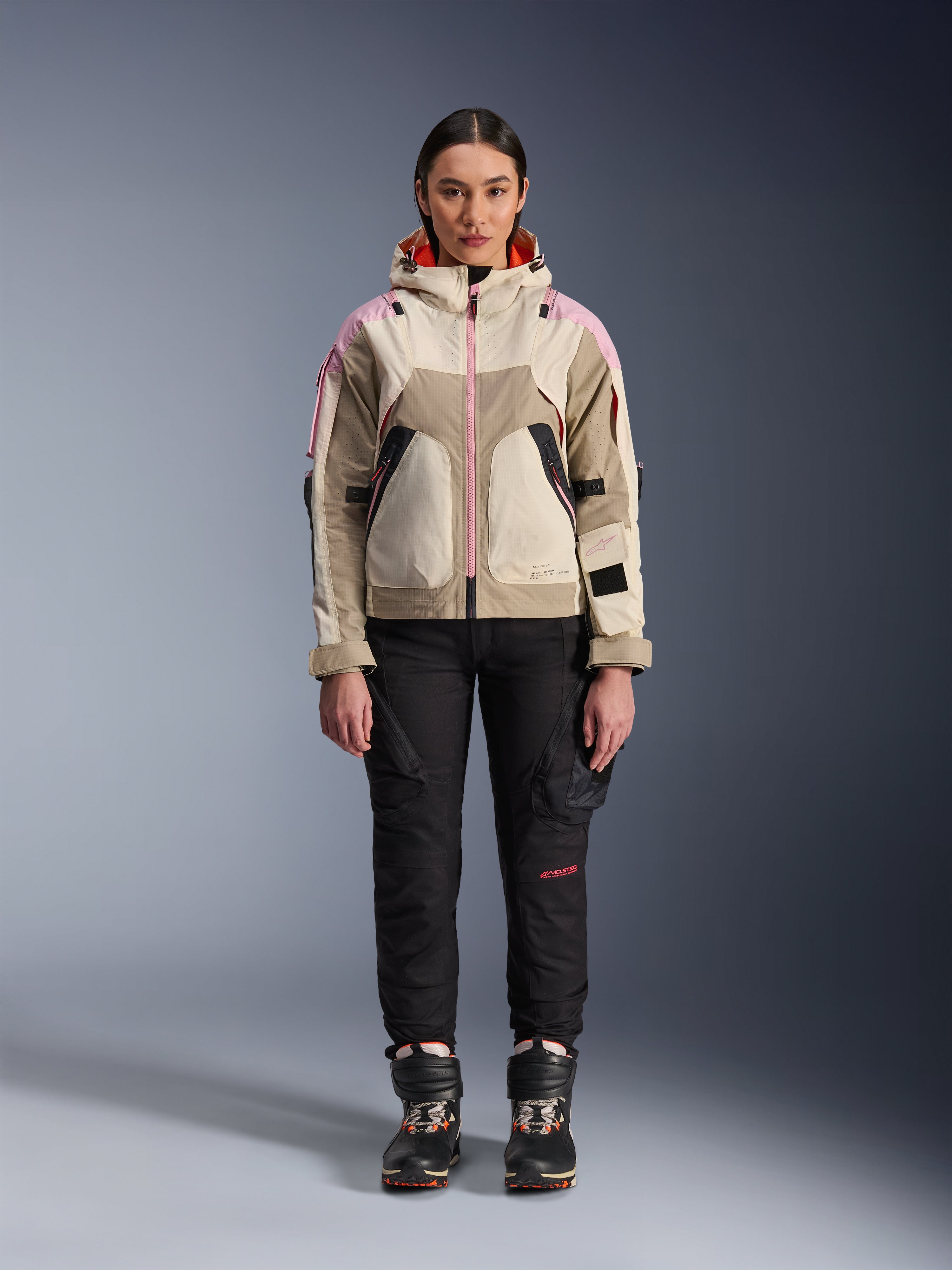 Modella che indossa la giacca Alpinestars Molly Air in Sand Pink Almond, abbinata a pantaloni tecnici da moto e stivali neri, vista frontale a figura intera, giacca da moto urbana con design d'ispirazione utility e accenti rosa