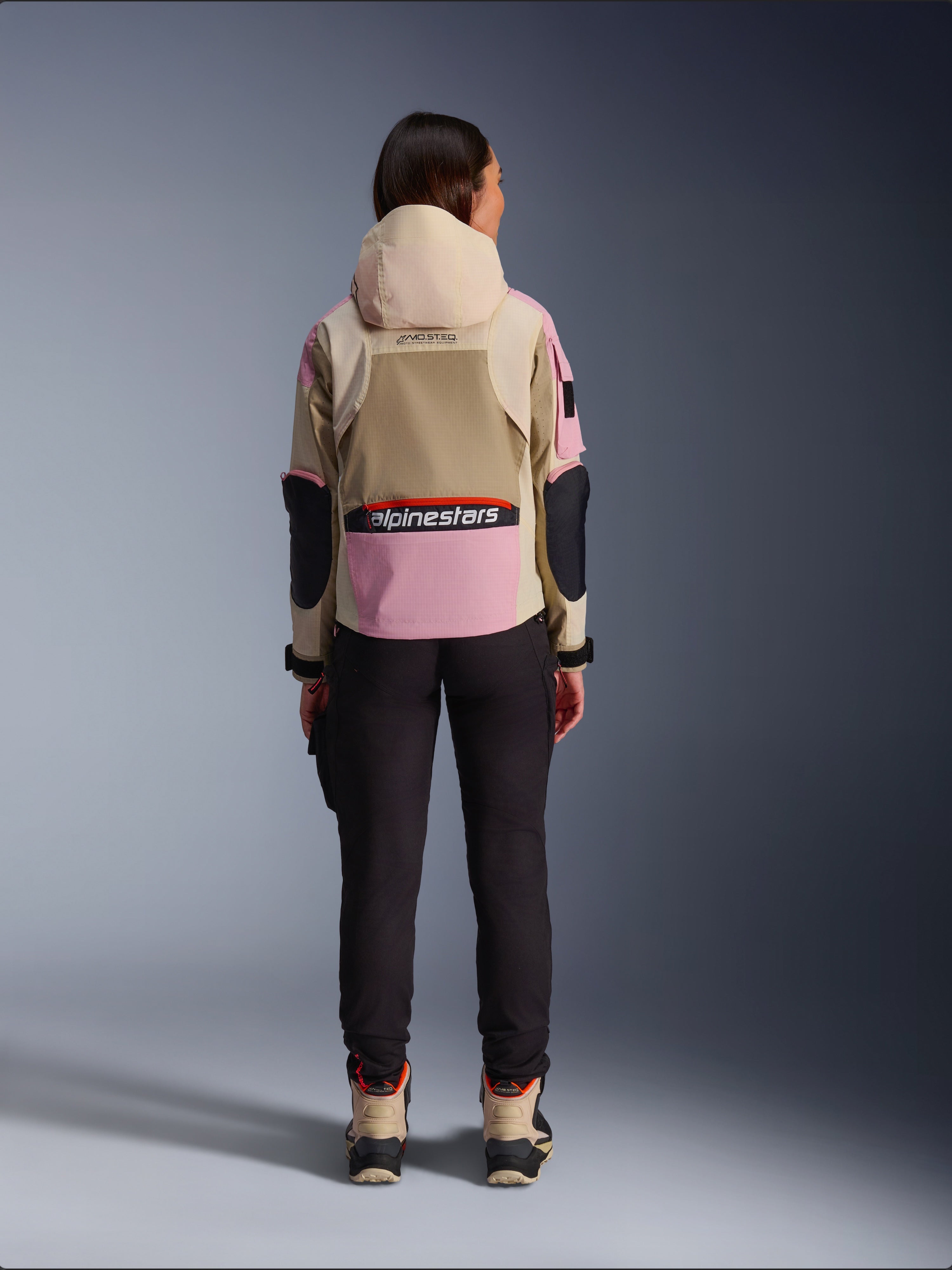 Motociclista vista da dietro che indossa la giacca Alpinestars Molly Air in Sand Pink Almond, con cappuccio beige e pannelli rosa con dettagli neri, abbinata a pantaloni da moto neri e stivali beige, abbigliamento da moto urban contemporaneo