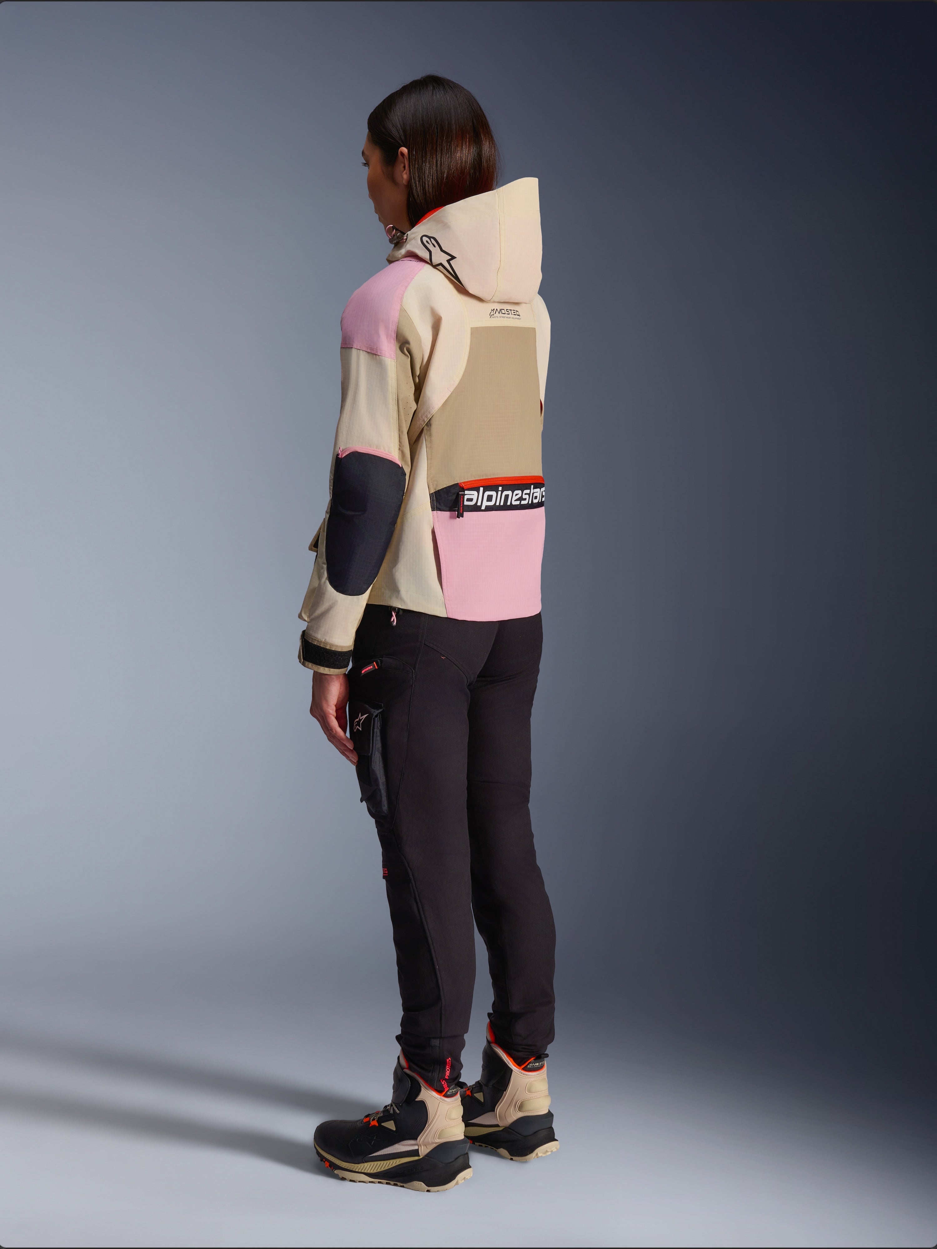 Motociclista che indossa la giacca Alpinestars Molly Air in Sand Pink Almond, caratterizzata da un design multi-tonale beige e rosa, mostrata da una vista laterale posteriore con pantaloni da moto neri e stivali tecnici abbinati, stile motociclistico urbano con cappuccio.