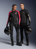 Uomo e donna in posa con abbigliamento da moto Alpinestars; uomo con giacca T-Dyno Air nera, donna con giacca in tessuto nera con accenti rosa e rossi, entrambi con caschi integrali neri, abbinati a pantaloni e stivali da moto neri.
