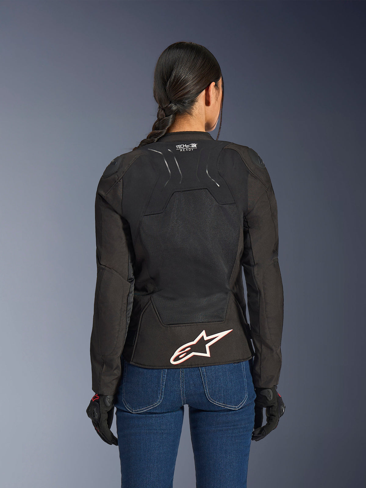 Giacca Alpinestars Stella SMX Air, vista posteriore su modella, tessuto nero con pannelli in mesh ad alto flusso, marchio 'TECH-AIR READY' sulla parte superiore della schiena, grande logo Alpinestars bianco con profilo rosso sulla parte inferiore della schiena, abbinata a jeans in denim blu e guanti da moto neri