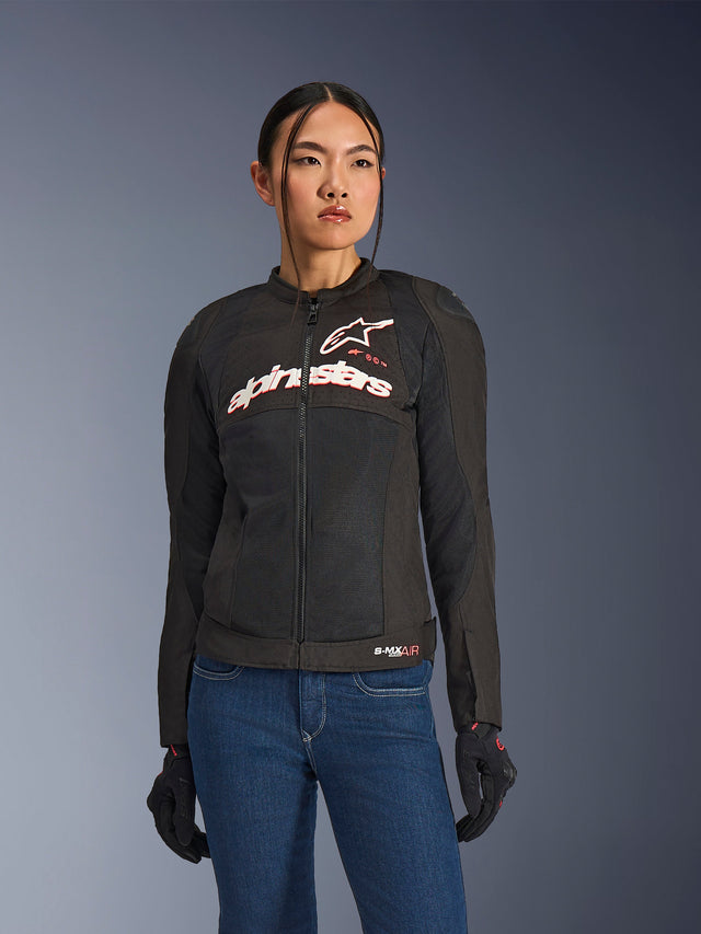Donna che indossa la giacca Alpinestars Stella SMX Air, nero bianco rosso fluo, giacca da moto in tessuto con rete ventilata e logo bianco sul petto, abbinata a jeans blu e guanti da guida neri, vestibilità femminile per la guida sportiva