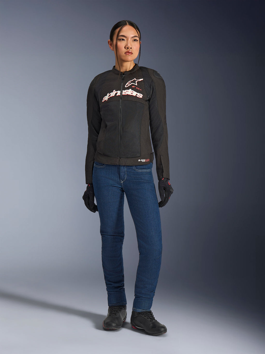 Motociclista con giacca Alpinestars Stella SMX Air, nera con loghi bianchi e rosso fluo, abbinata a pantaloni da moto in denim blu e guanti neri, giacca sportiva in tessuto ventilato per uso stradale