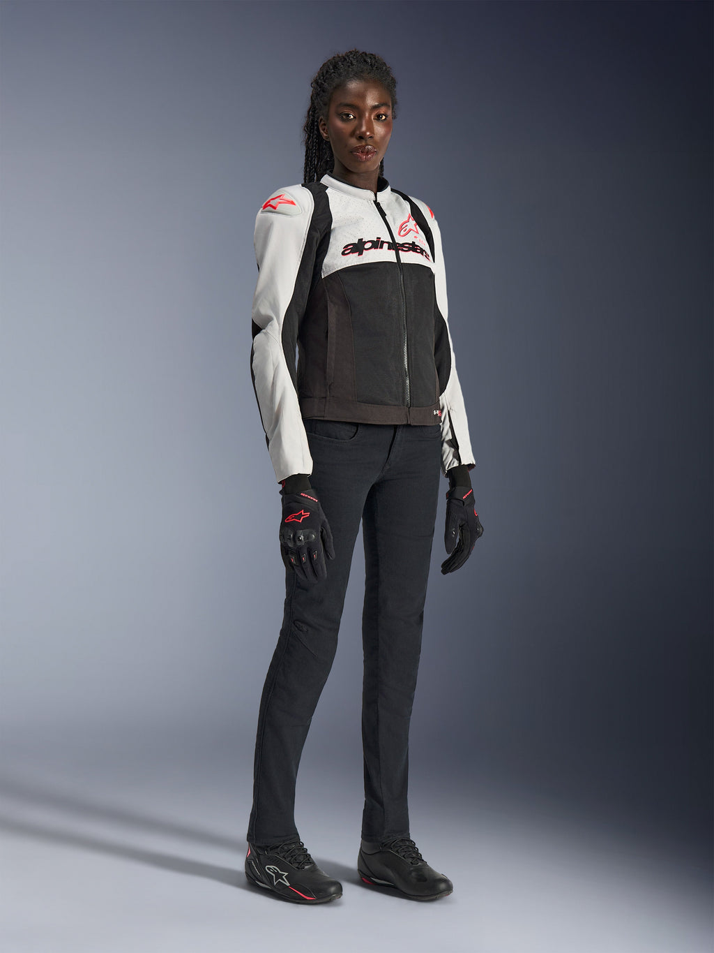 Giacca Alpinestars Stella SMX Air da donna in grigio ghiaccio, nero e corallo fluo, indossata da una modella con pantaloni da moto e guanti neri, caratterizzata da struttura in rete e loghi rosso corallo, abbigliamento tecnico sportivo da donna