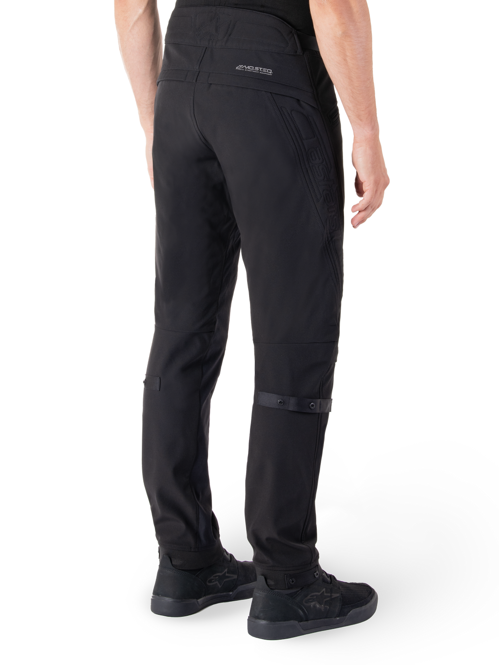 Alpinestars Alden Pants, pantaloni da moto urban, nero, vista posteriore che mostra il tessuto softshell tecnico con dettagli trapuntati, branding riflettente MO.ST.EQ. sul carré posteriore e cinturini regolabili sui polpacci, abbinati a scarpe da guida nere
