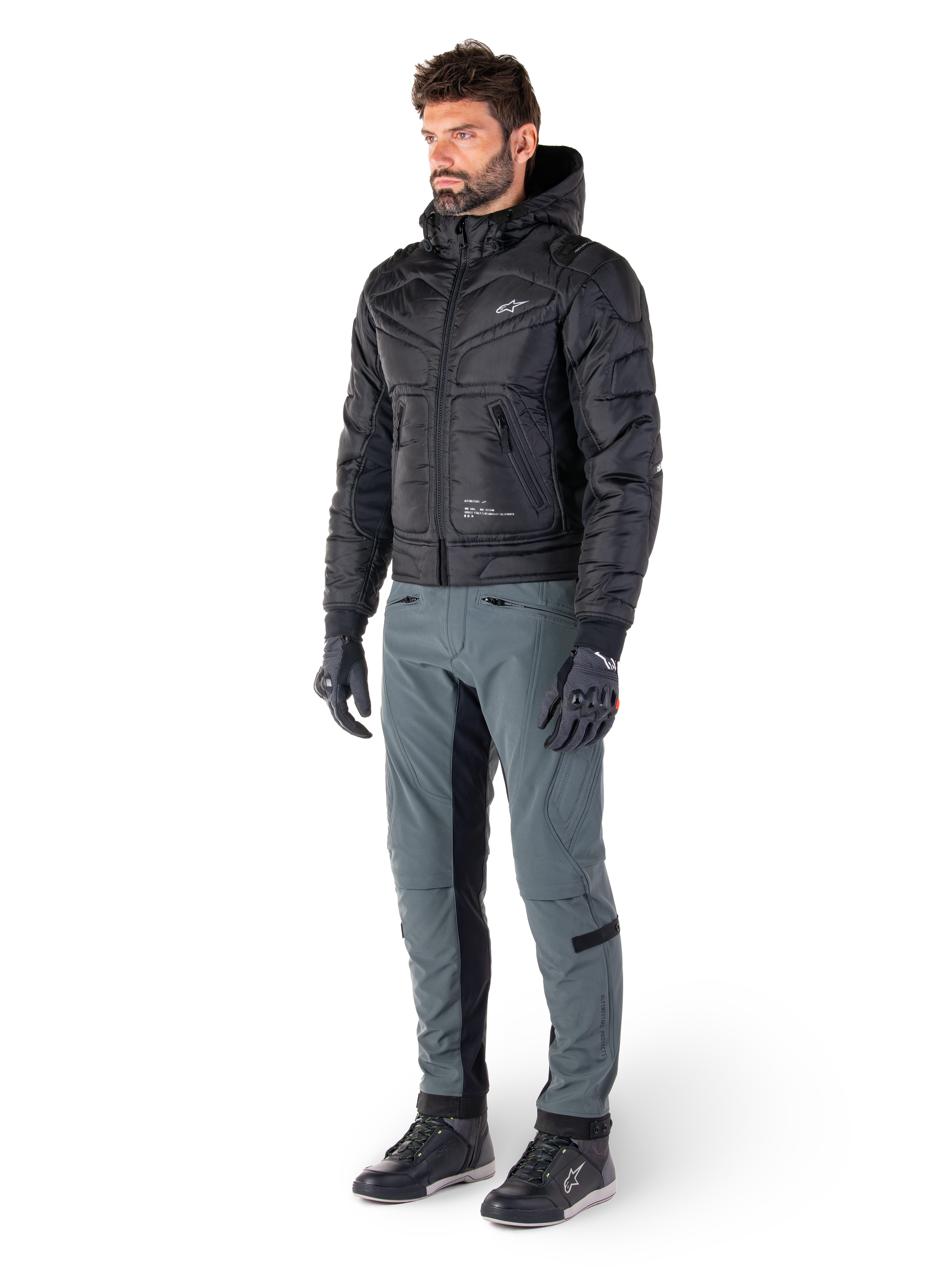 Pantaloni Alpinestars Alden in Urban Gray, indossati da un modello con giacca nera trapuntata con cappuccio, guanti e scarpe da moto, con dettagli trapuntati sui lati e pannelli neri sull'interno gamba, abbigliamento da moto urbano d'ispirazione streetwear