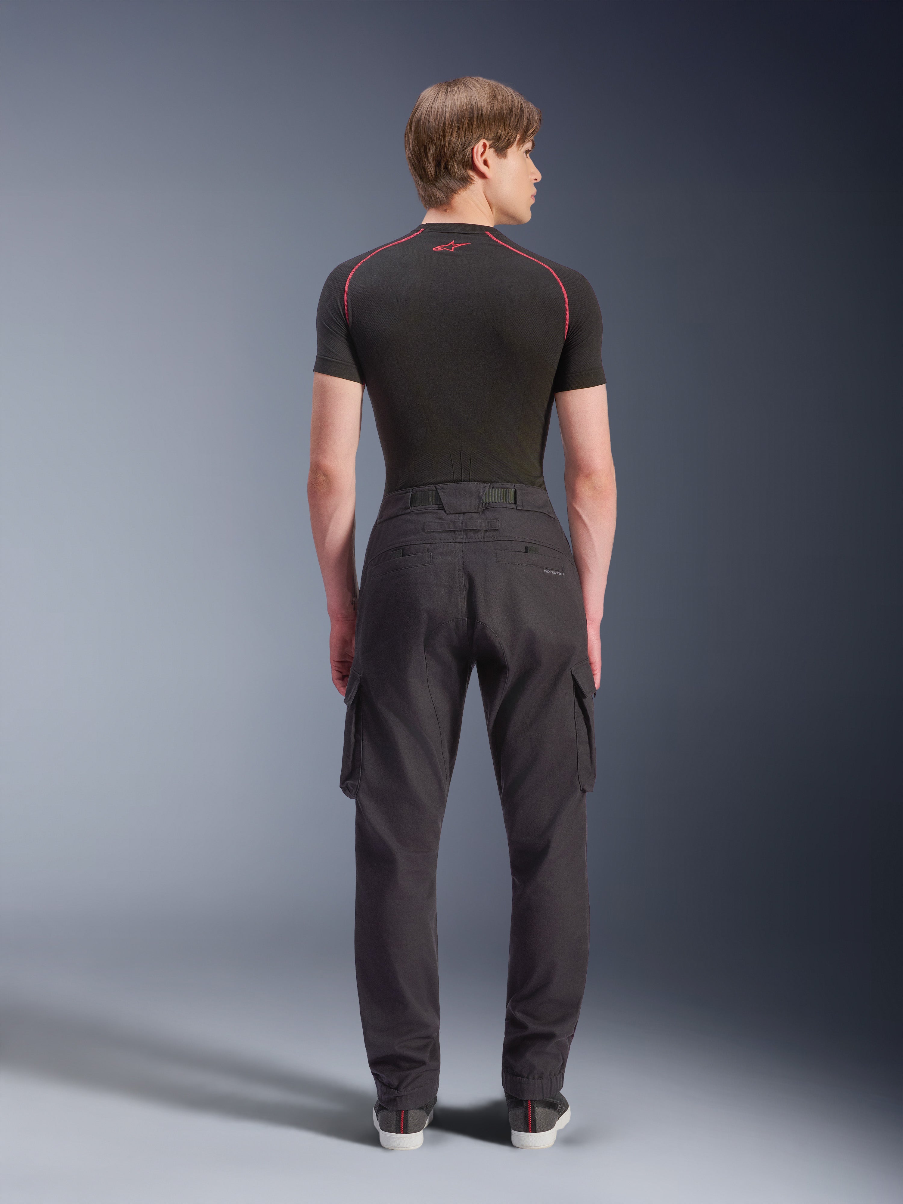 Alpinestars Flex-AST Cargo Pants, pantaloni cargo da moto, nero, vista posteriore su modello, con vestibilità regular tapered, orli elasticizzati e tasche cargo laterali, abbinati a un base layer nero e sneaker casual