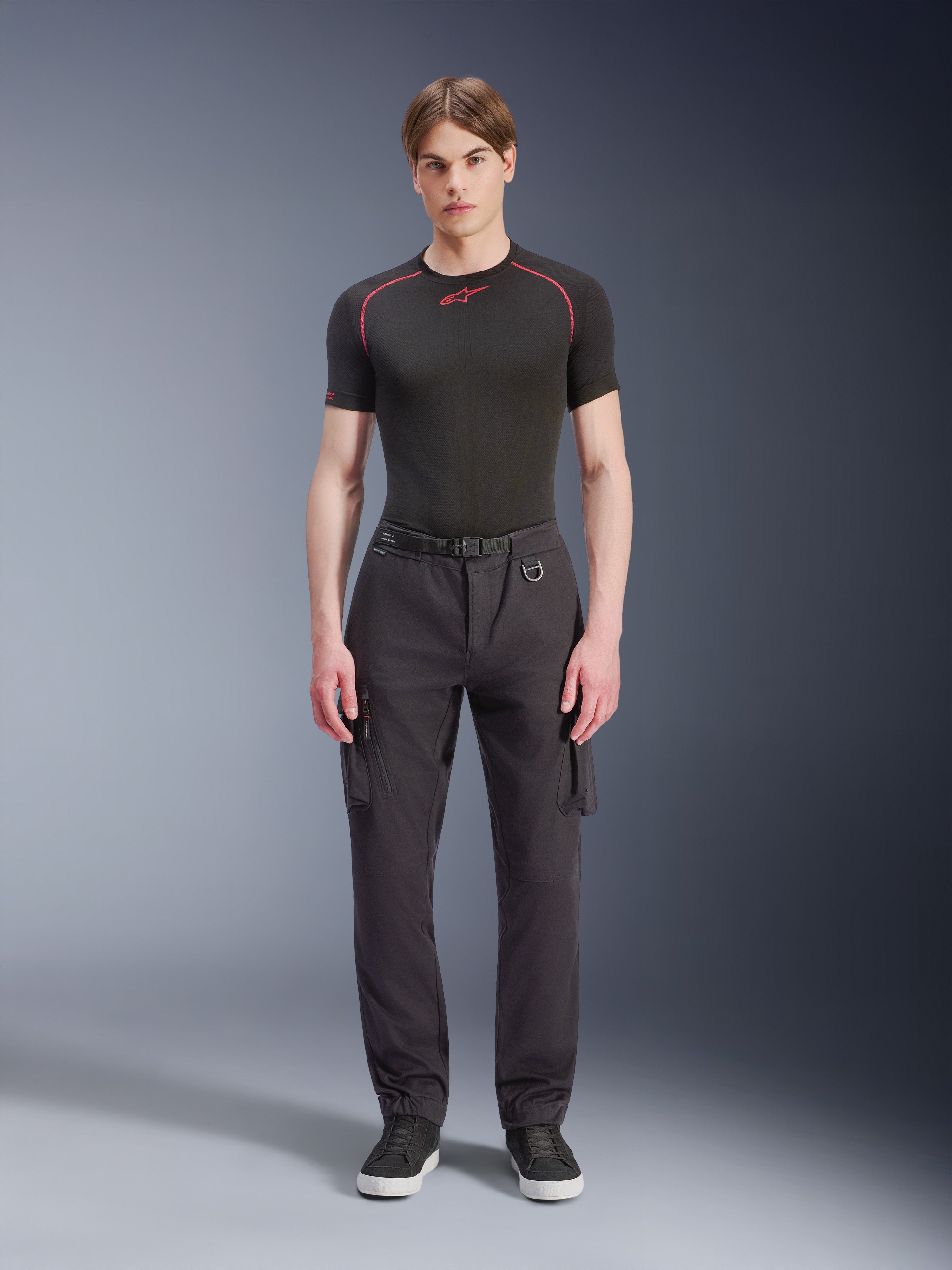 Alpinestars Pantaloni Cargo Flex-AST - Corti, nero, indossati da un modello con una t-shirt tecnica nera e sneakers nere, pantaloni da moto urbani con diverse tasche cargo, vestibilità affusolata e cintura integrata con fibbia e anello a D