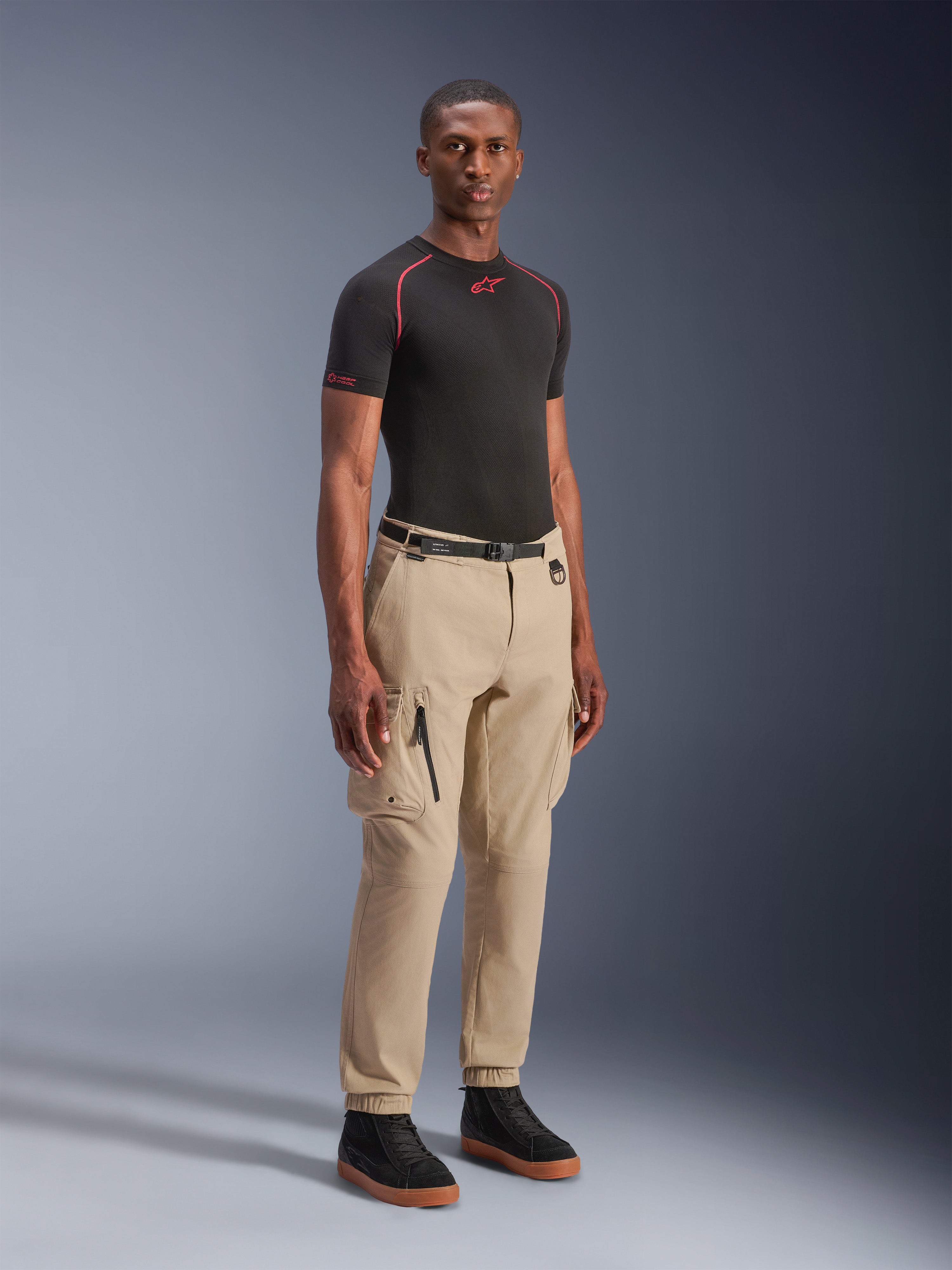 Modello che indossa i pantaloni cargo Alpinestars Flex-AST in Light Beige, vestibilità regular affusolata con orli elasticizzati, ampie tasche cargo sulle cosce, cintura nera in vita, abbinati a un base layer nero a maniche corte e sneakers nere, stile motociclistico urbano