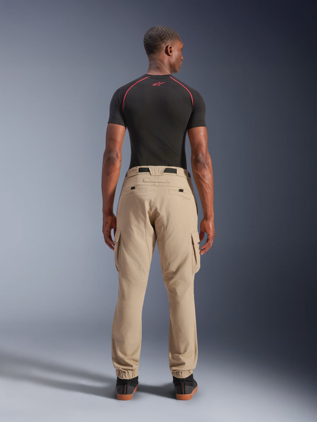 Alpinestars Pantaloni Cargo Flex-AST Short, beige chiaro, vista posteriore su un modello, vestibilità regolare affusolata con orli elasticizzati e tasche cargo, indossati con base layer tecnico nero con accenti rossi, abbigliamento moto urbano moderno
