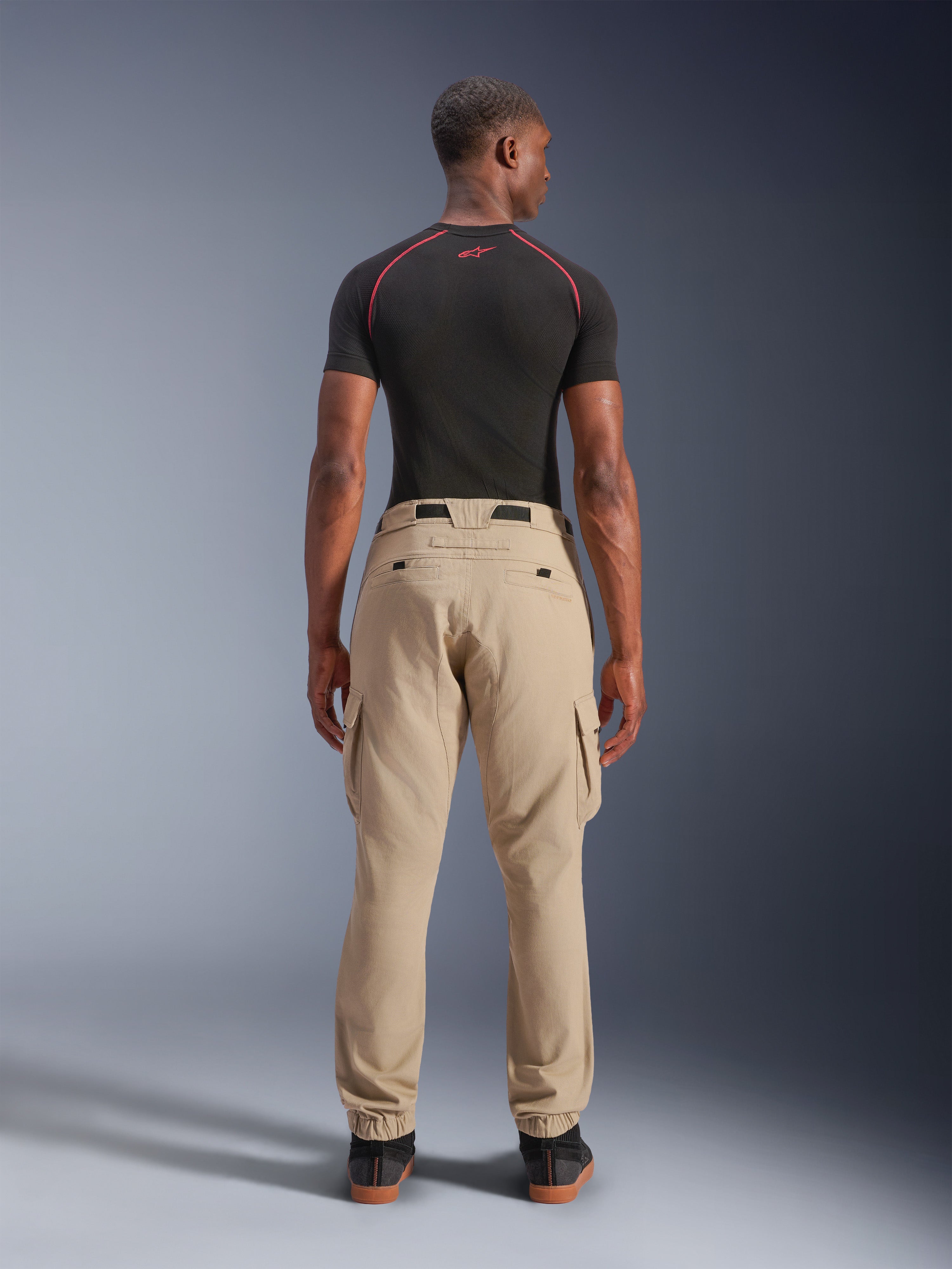 Alpinestars Pantaloni Cargo Flex-AST Short, beige chiaro, vista posteriore su un modello, vestibilità regolare affusolata con orli elasticizzati e tasche cargo, indossati con base layer tecnico nero con accenti rossi, abbigliamento moto urbano moderno