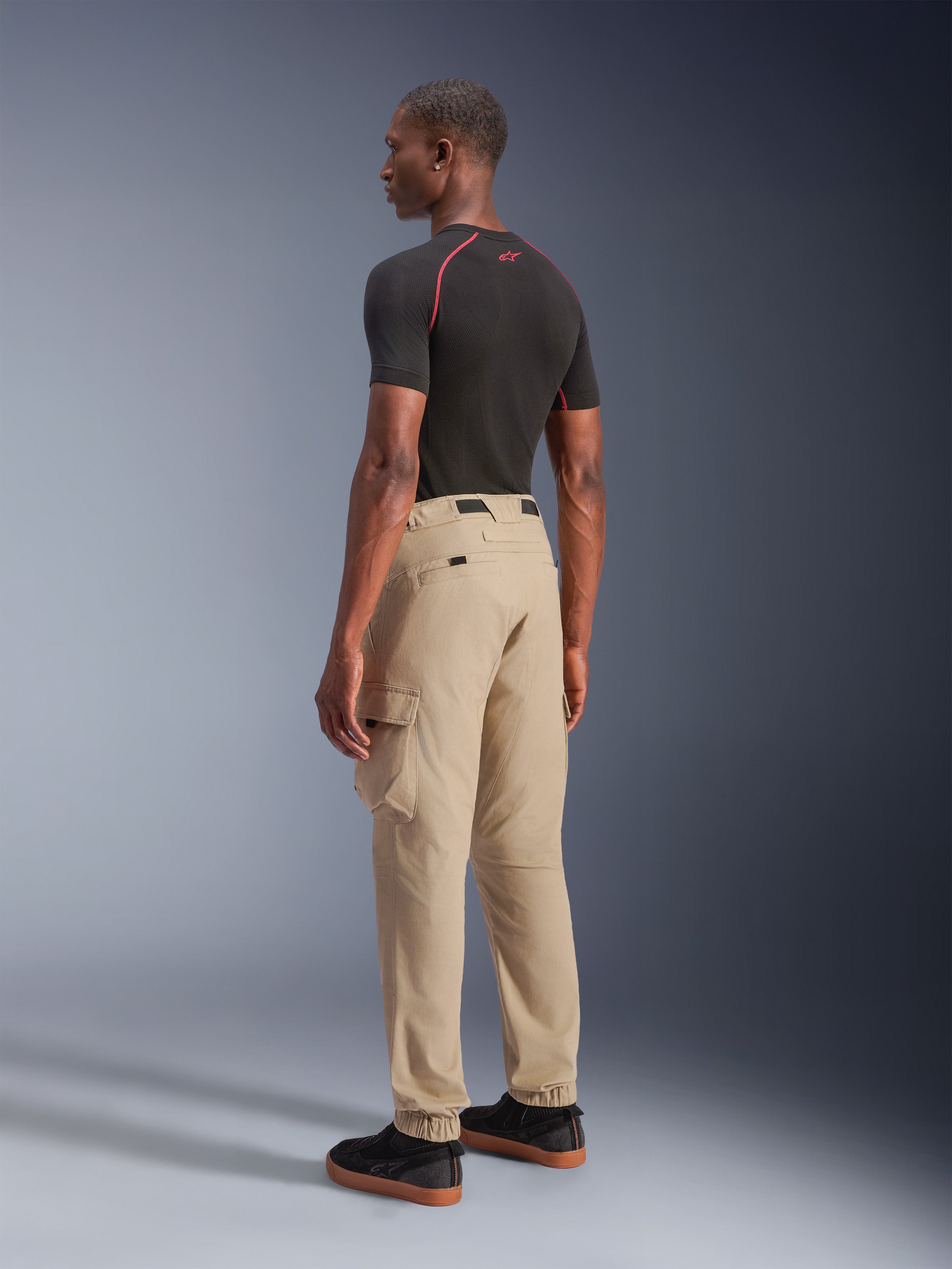 Alpinestars Flex-AST Cargo Pants - Short, pantaloni da moto urban in beige chiaro, vista posteriore di tre quarti con tasche cargo laterali e orli elasticizzati, abbinati a una maglia nera a maniche corte e scarpe casual nere