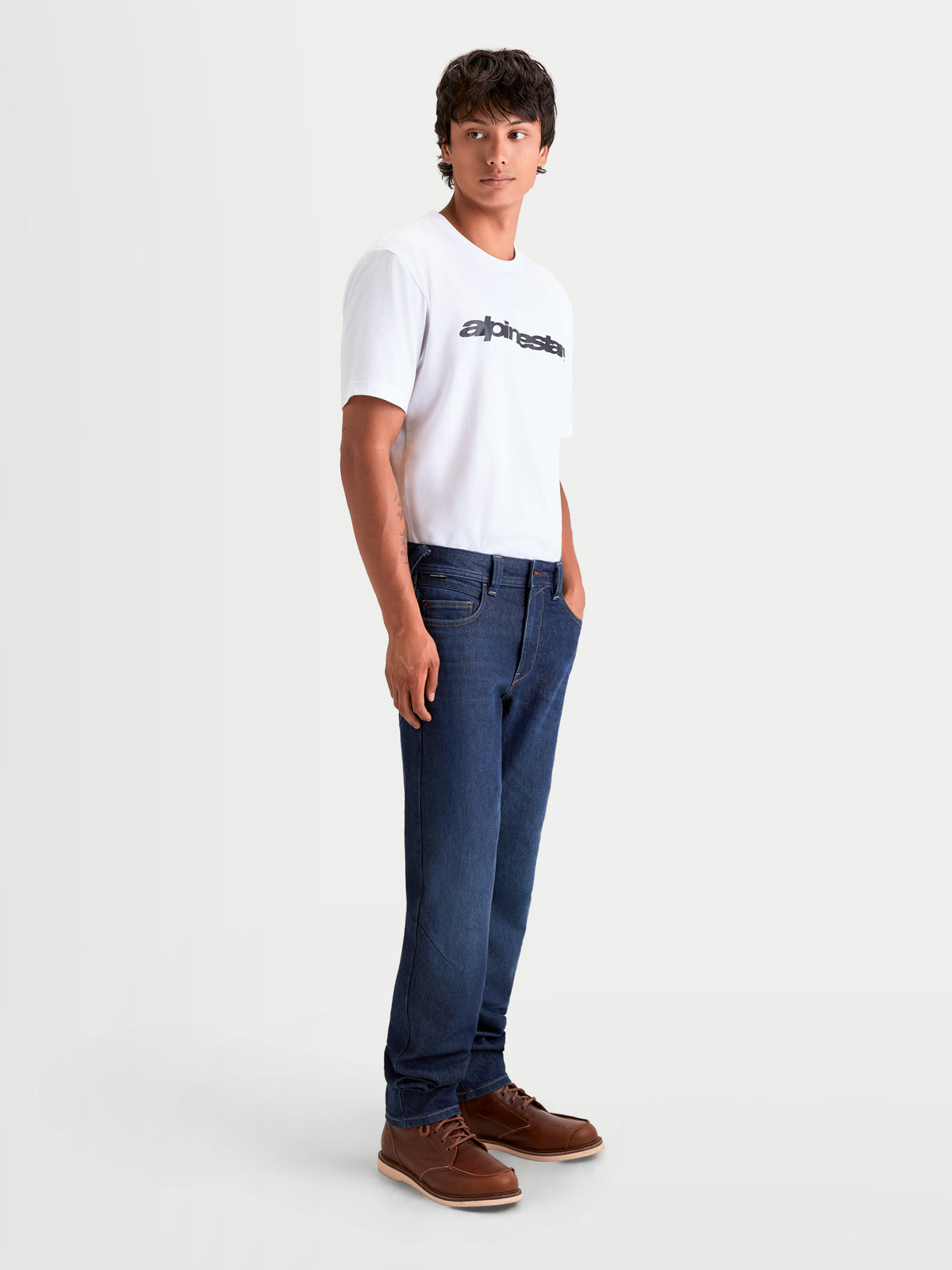Alpinestars Flex Ast 7 Denim Pantaloni, jeans da moto urbani da uomo in blu scuro, vista laterale indossati da un modello con t-shirt bianca e stivali marroni, con struttura in CORDURA® a strato singolo certificata AA