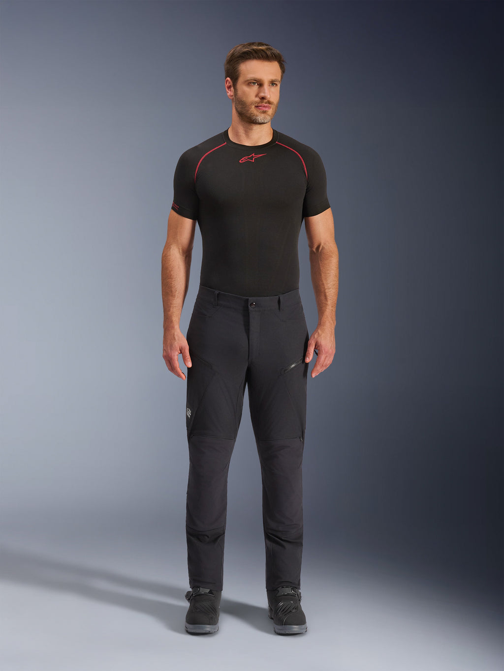 Alpinestars Flex-AST Explorer Pants, pantaloni da moto adventure touring neri, uomo che indossa pantaloni tecnici aderenti con tasche cargo con zip e pannelli rinforzati, abbinati a base layer nero e stivali da moto, protezione resistente per la guida adventure