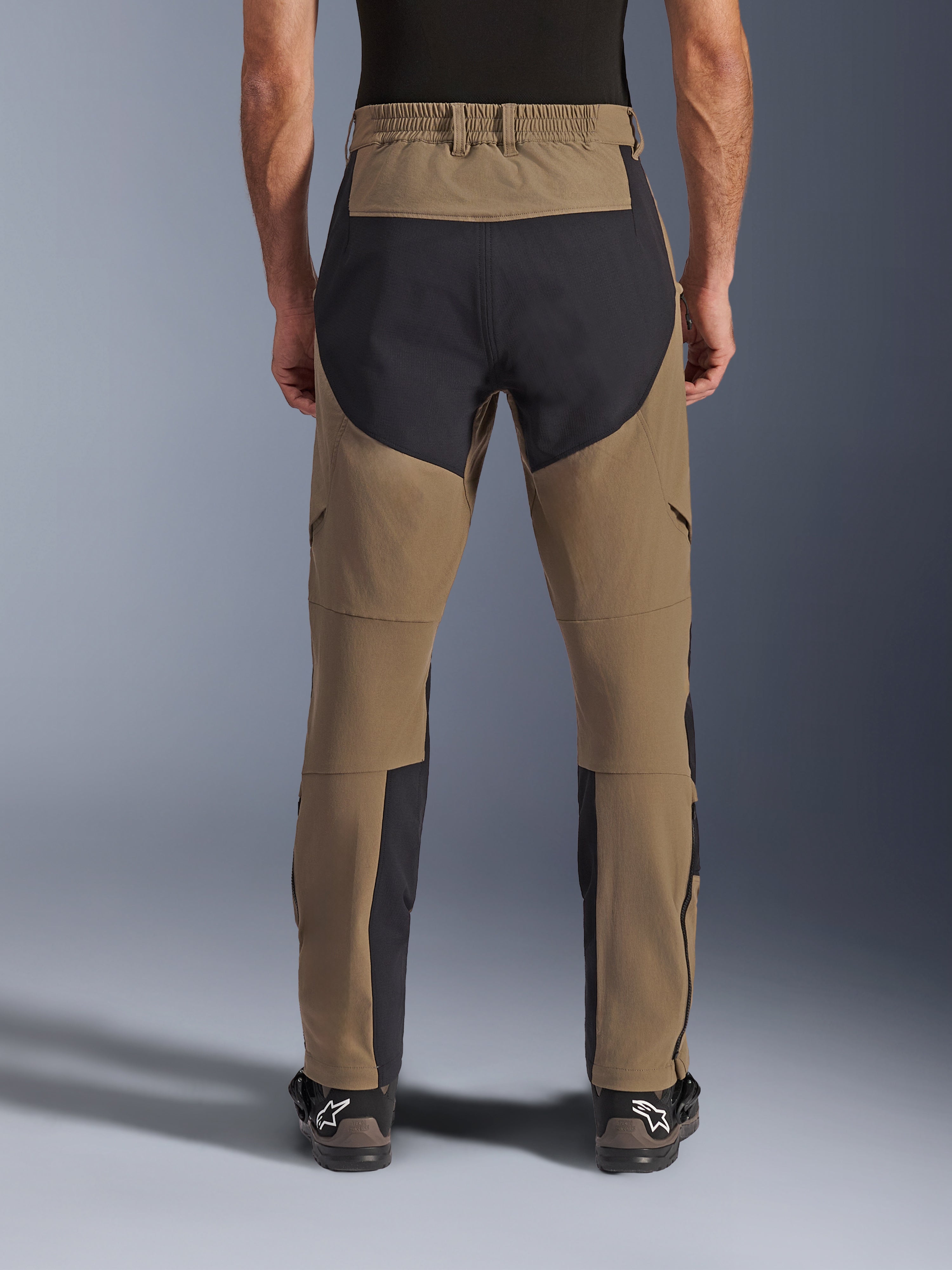 Pantaloni Alpinestars Flex-AST Explorer, pantaloni da moto adventure, Walnut Black, vista posteriore indossata dal pilota, pannelli di rinforzo in ripstop nero sulla seduta e sull'interno gamba, vita elasticizzata, lunghe cerniere sulla parte inferiore della gamba e stivali adventure coordinati