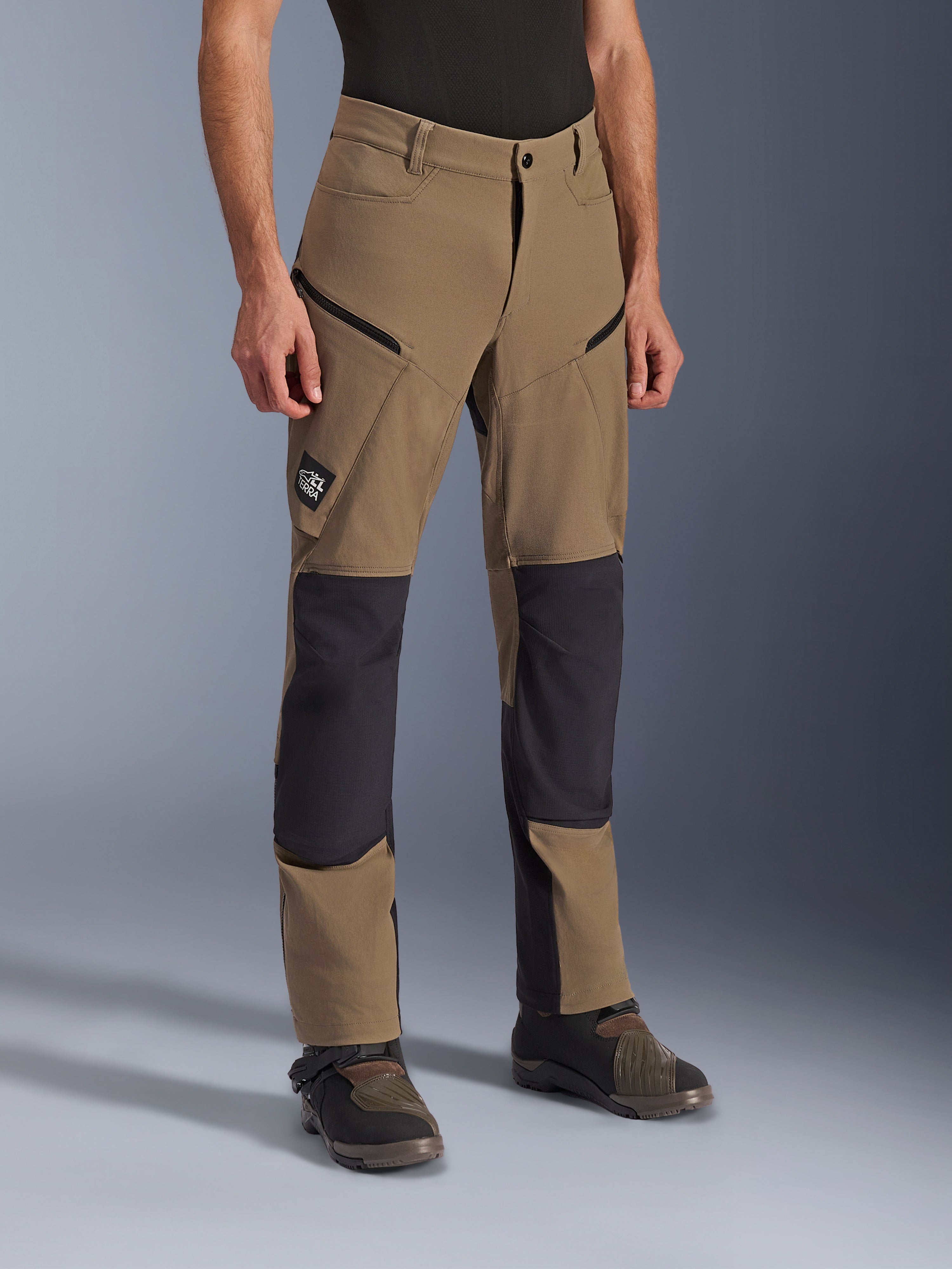 Alpinestars Flex-AST Explorer Pants, pantaloni da moto adventure in Walnut Black, in tessuto color sabbia con pannelli neri rinforzati su ginocchia e seduta, tasche cargo con cerniera e patch logo All Terra, mostrati indossati con stivali tecnici da guida.