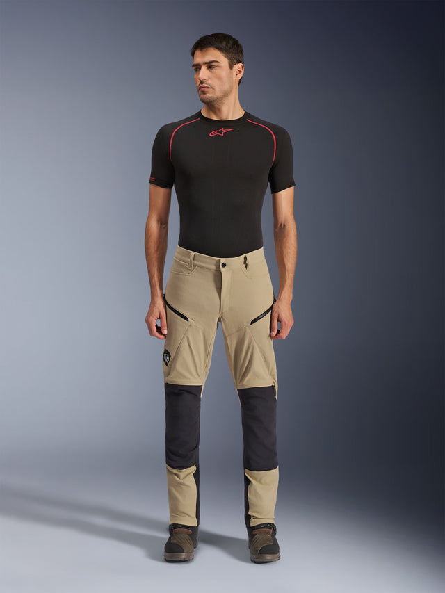 Alpinestars Flex-AST Explorer Pants, pantaloni da moto adventure, beige e nero, con pannelli rinforzati sulle ginocchia, tasche cargo con zip e costruzione in tessuto ripstop, indossati da un modello in uno scatto in studio frontale.