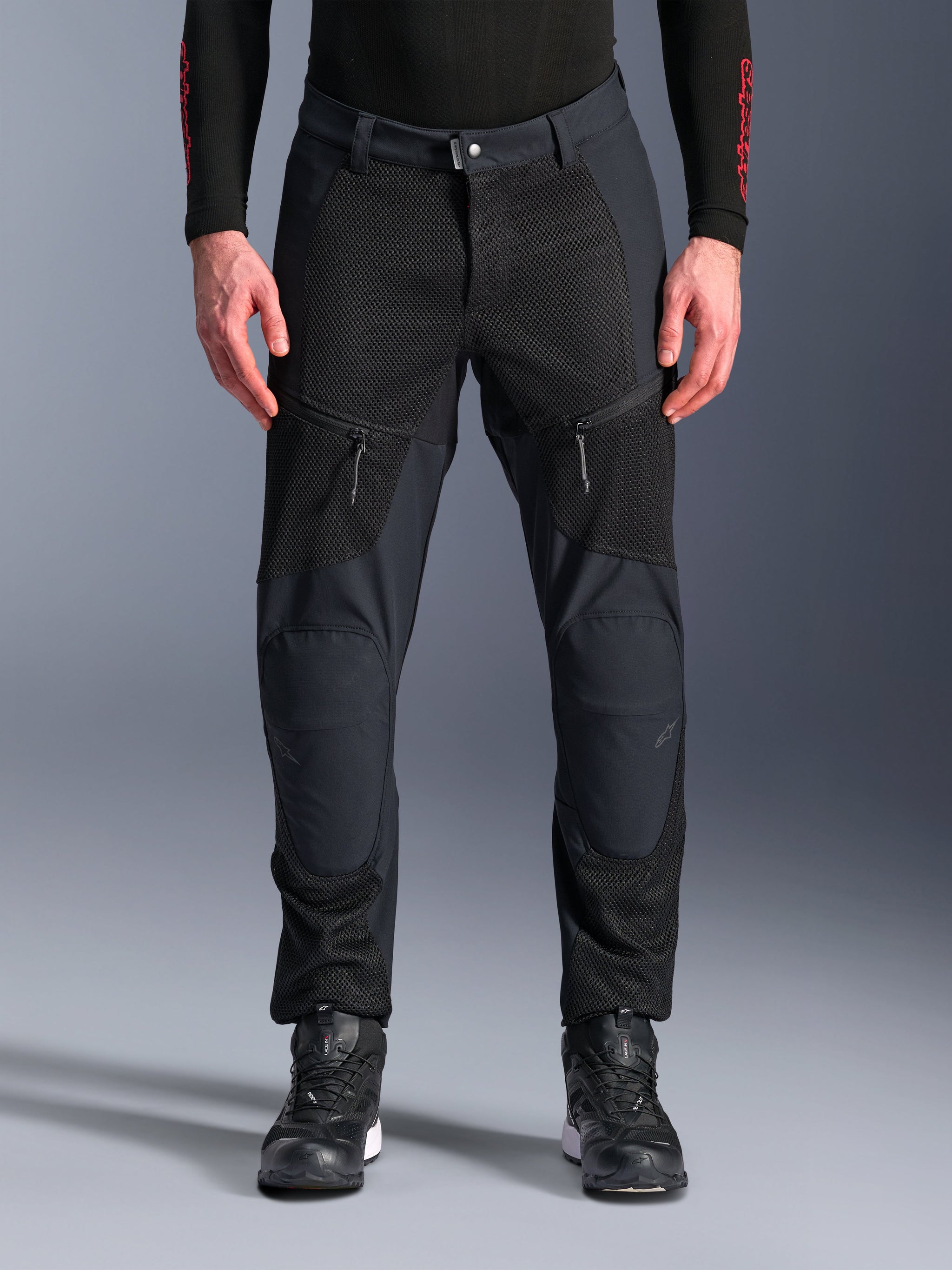 Alpinestars Troop-Air Pro Pants, pantaloni da moto neri, con ampi pannelli di ventilazione in rete sulle cosce e sulla parte inferiore delle gambe, tasche anteriori con zip, design in tessuto con vestibilità sportiva, indossati con scarpe da moto nere e un base layer a maniche lunghe