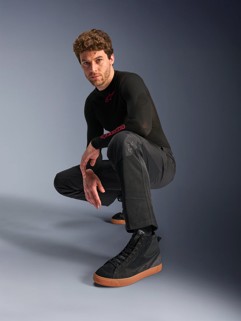 Alpinestars C-1 Air Pants, pantaloni da moto urban, nero, modello accovacciato in studio, indossati con maglia nera a maniche lunghe e sneakers nere con suole marroni, dotati di pannelli in mesh traspirante e stile urban contemporaneo