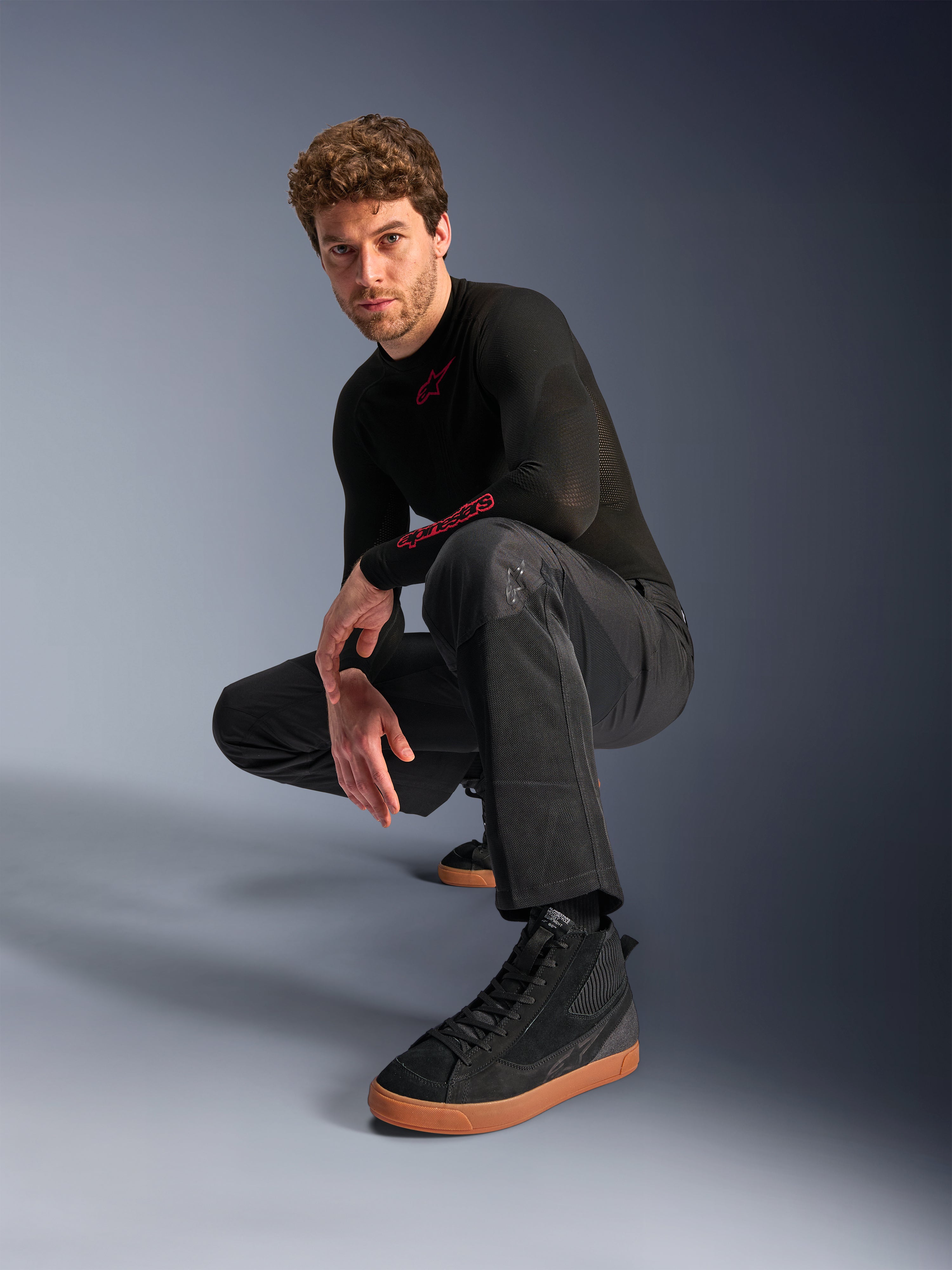 Alpinestars C-1 Air Pants, pantaloni da moto urban, nero, modello accovacciato in studio, indossati con maglia nera a maniche lunghe e sneakers nere con suole marroni, dotati di pannelli in mesh traspirante e stile urban contemporaneo