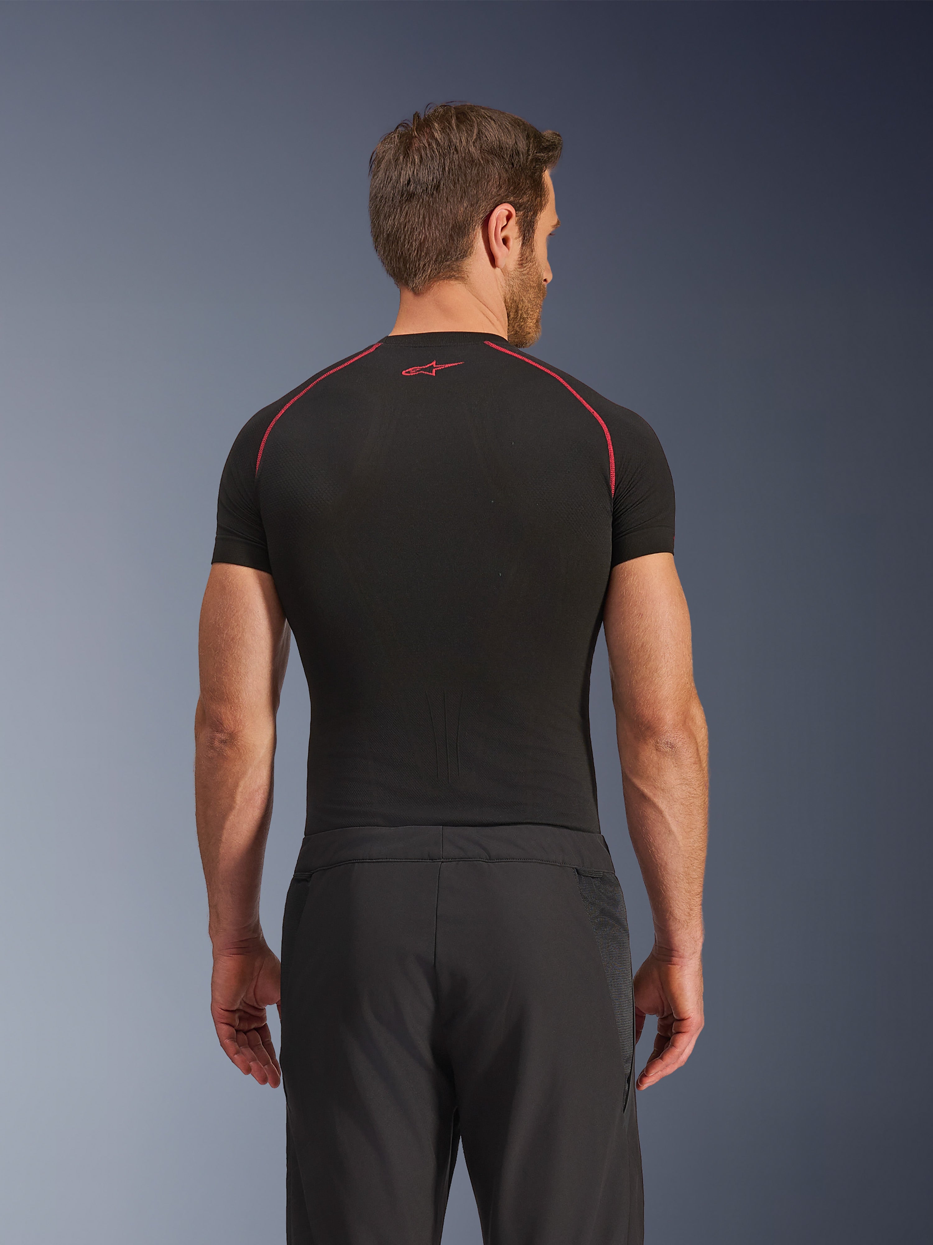 Vista posteriore di un modello che indossa i pantaloni Alpinestars Aeroshell Airflow, neri, costruzione leggera in softshell con pannelli laterali in rete integrati, abbinati a un base layer nero con cuciture rosse a contrasto, abbigliamento sportivo da moto