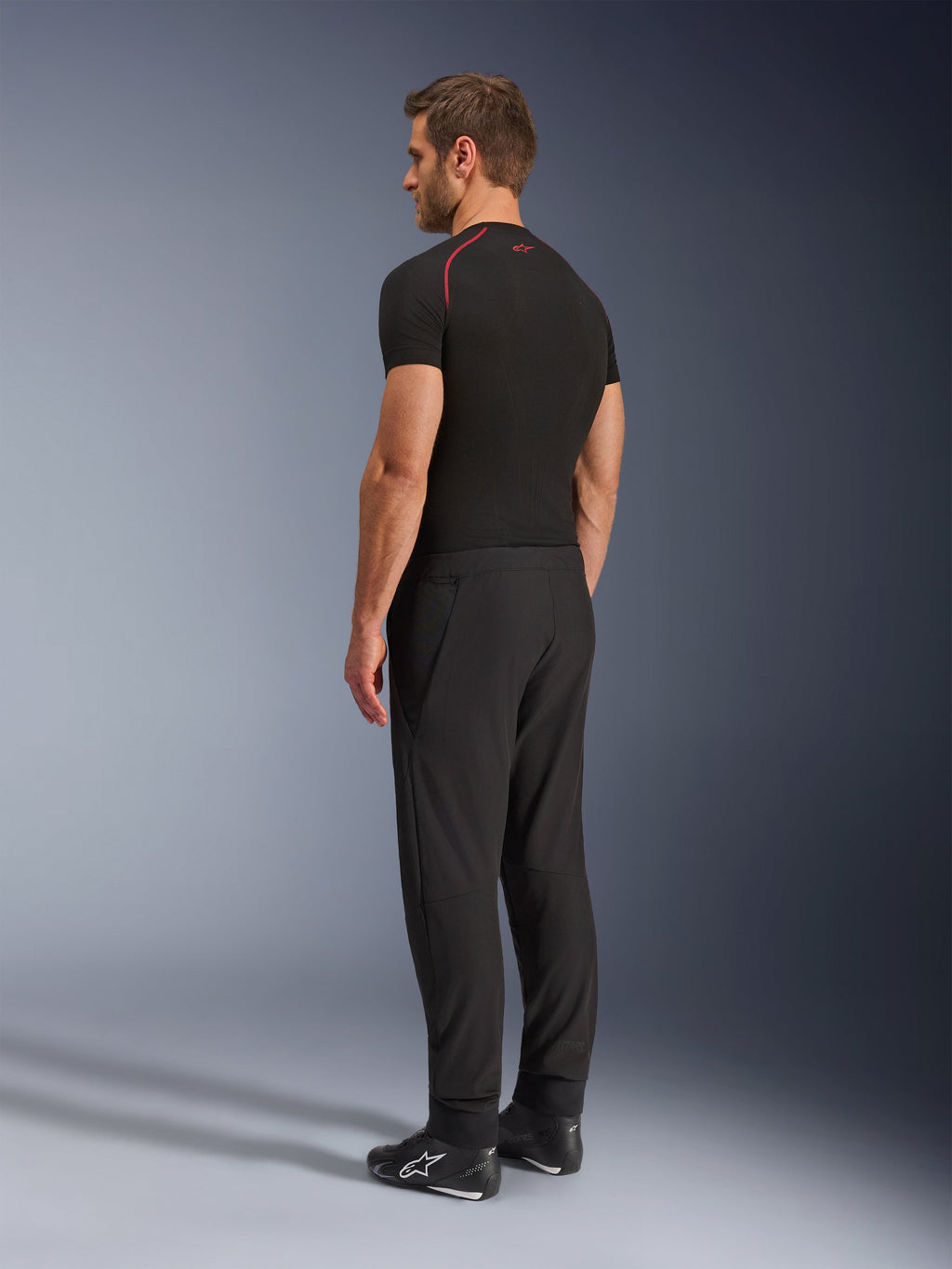Pantaloni Alpinestars Aeroshell Airflow, pantaloni da moto neri, leggero tessuto softshell, indossati con maglia tecnica nera a maniche corte e stivali da moto neri, vista laterale posteriore che mostra la vestibilità slim e la costruzione ergonomica