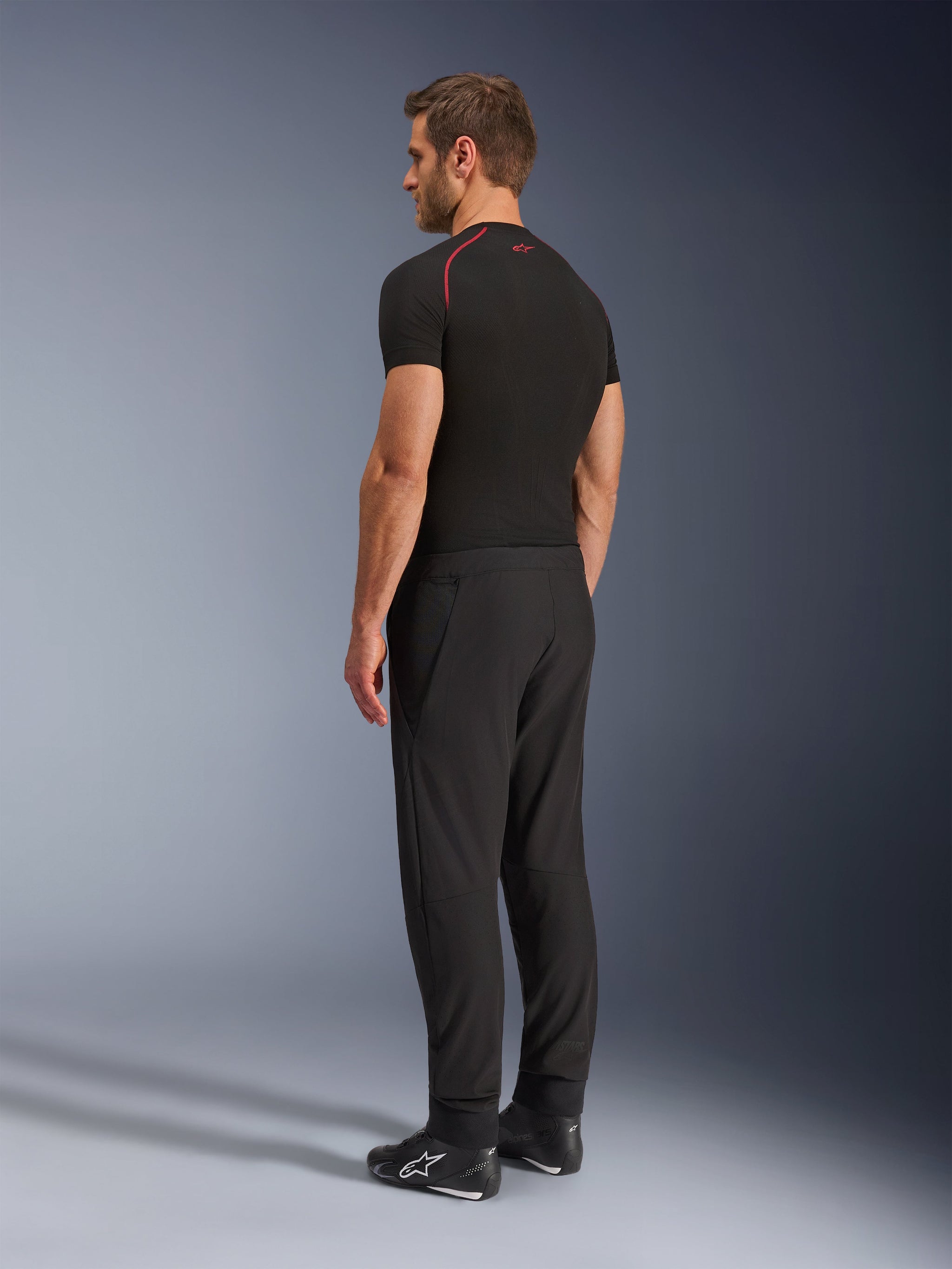 Pantaloni Alpinestars Aeroshell Airflow, pantaloni da moto neri, leggero tessuto softshell, indossati con maglia tecnica nera a maniche corte e stivali da moto neri, vista laterale posteriore che mostra la vestibilità slim e la costruzione ergonomica
