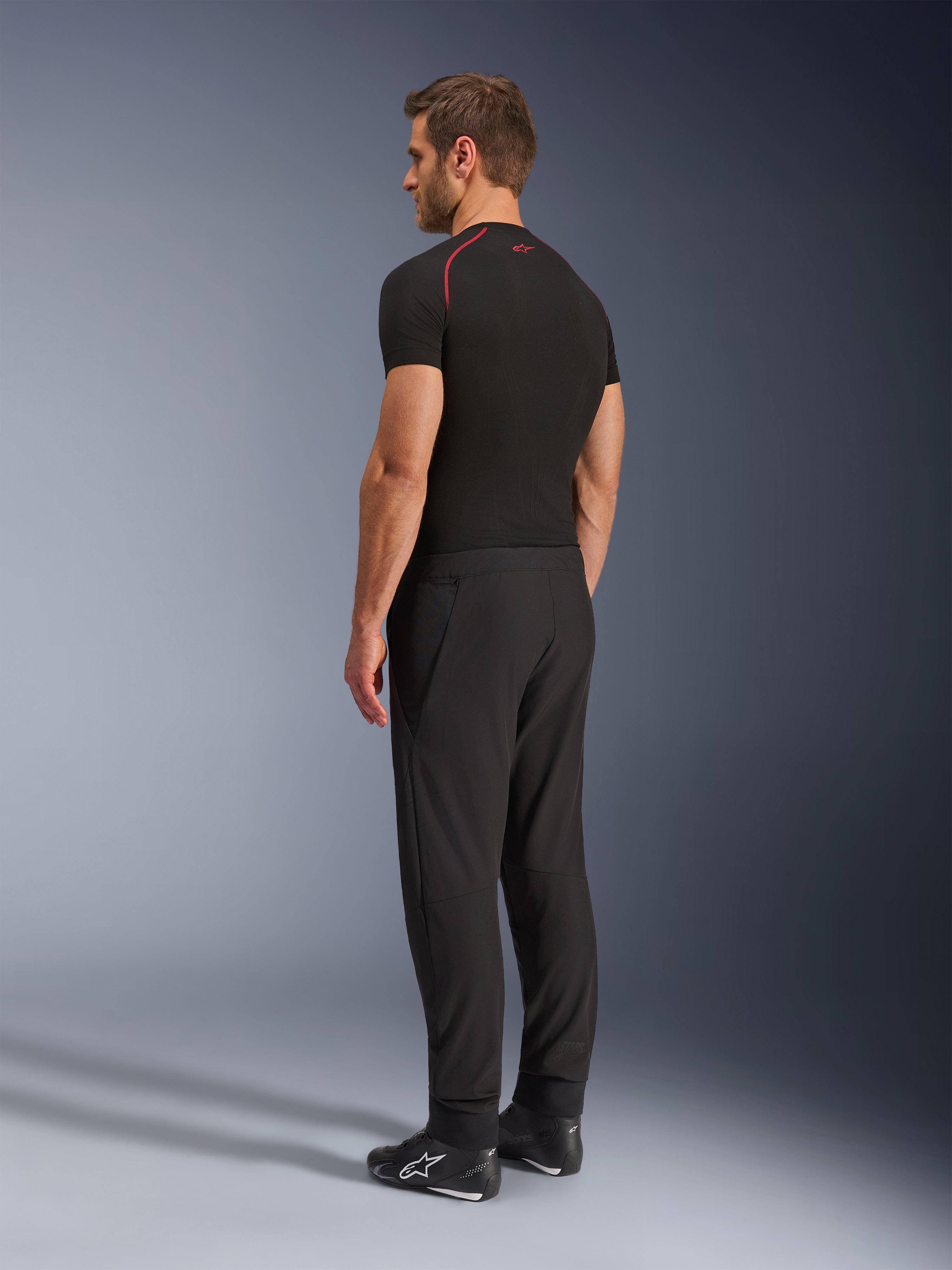 Pantaloni Alpinestars Aeroshell Airflow, pantaloni da moto neri, leggero tessuto softshell, indossati con maglia tecnica nera a maniche corte e stivali da moto neri, vista laterale posteriore che mostra la vestibilità slim e la costruzione ergonomica