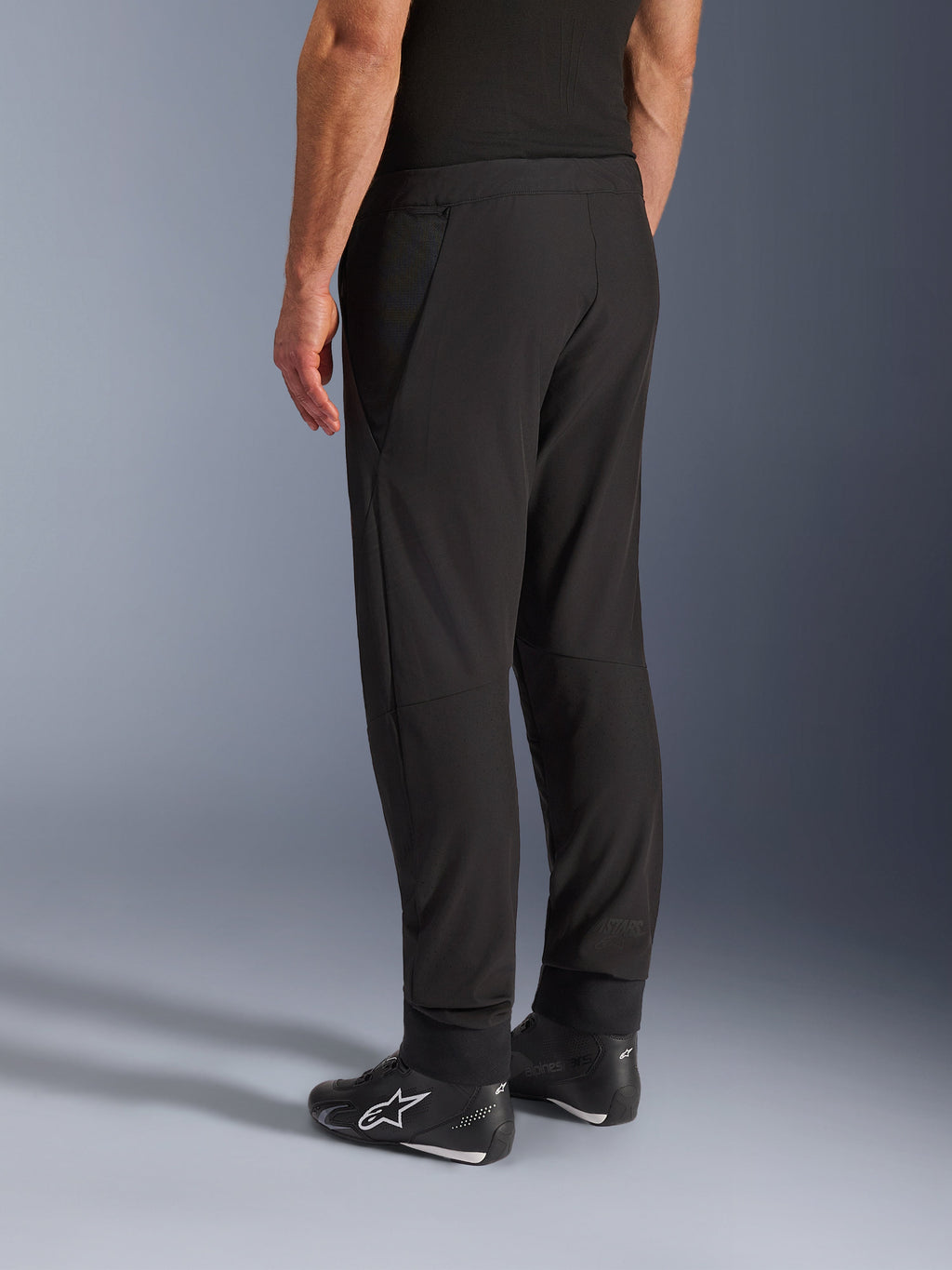 Pantaloni Alpinestars Aeroshell Airflow, pantaloni da moto neri, vista posteriore a tre quarti, costruzione in leggero softshell elasticizzato con pannelli in rete integrati, polsini delle caviglie regolabili, indossati con scarpe da moto Alpinestars nere