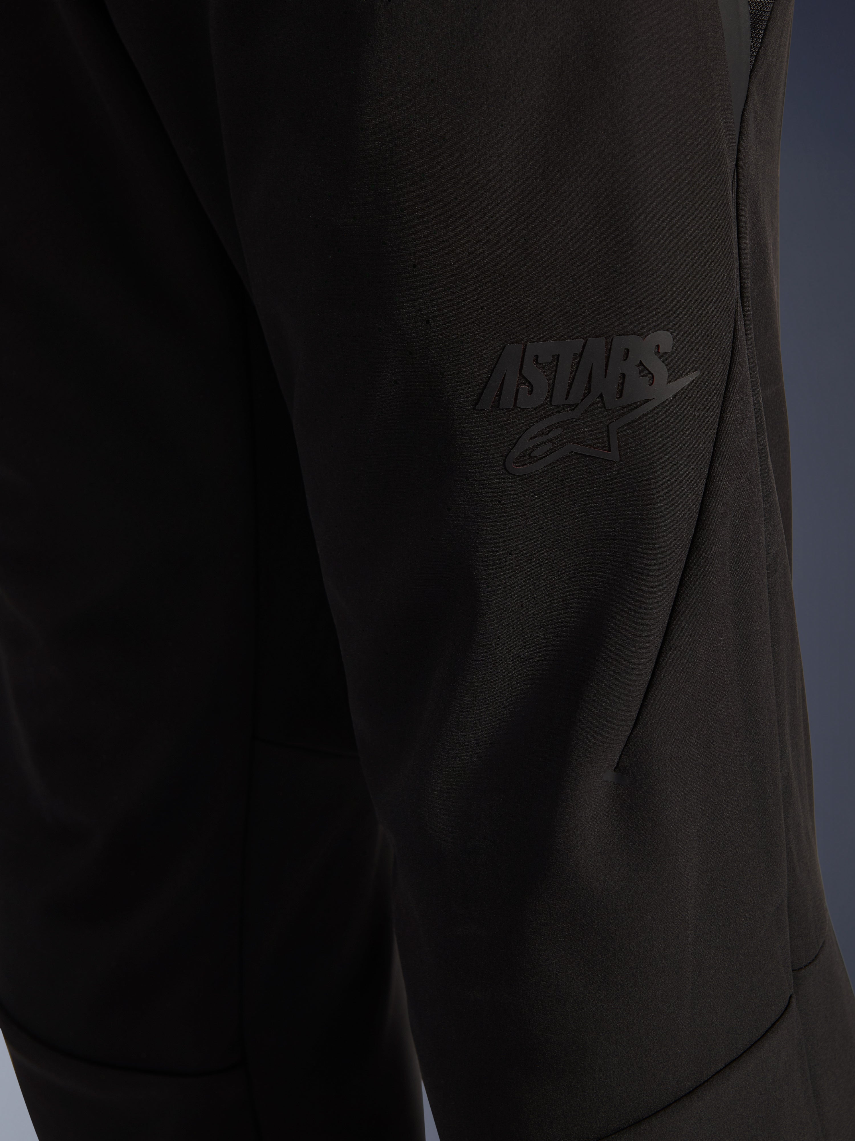 Alpinestars Aeroshell Airflow Pants, pantaloni da moto neri, vista ravvicinata della zona della coscia con logo ASTARS nero a rilievo, materiale softshell elasticizzato leggero con design ergonomico delle cuciture