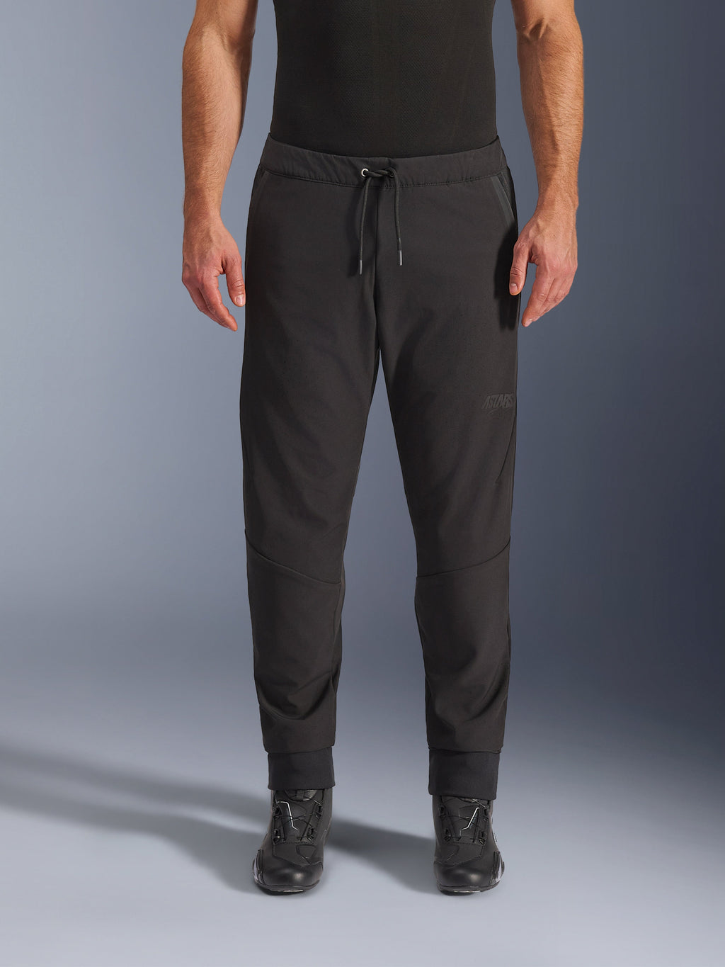 Pantaloni Alpinestars Aeroshell Airflow, pantaloni da moto neri, vista frontale indossati con stivali da moto neri, presentano vita con coulisse e polsini alle caviglie affusolati, design in softshell leggero con branding tonale discreto.