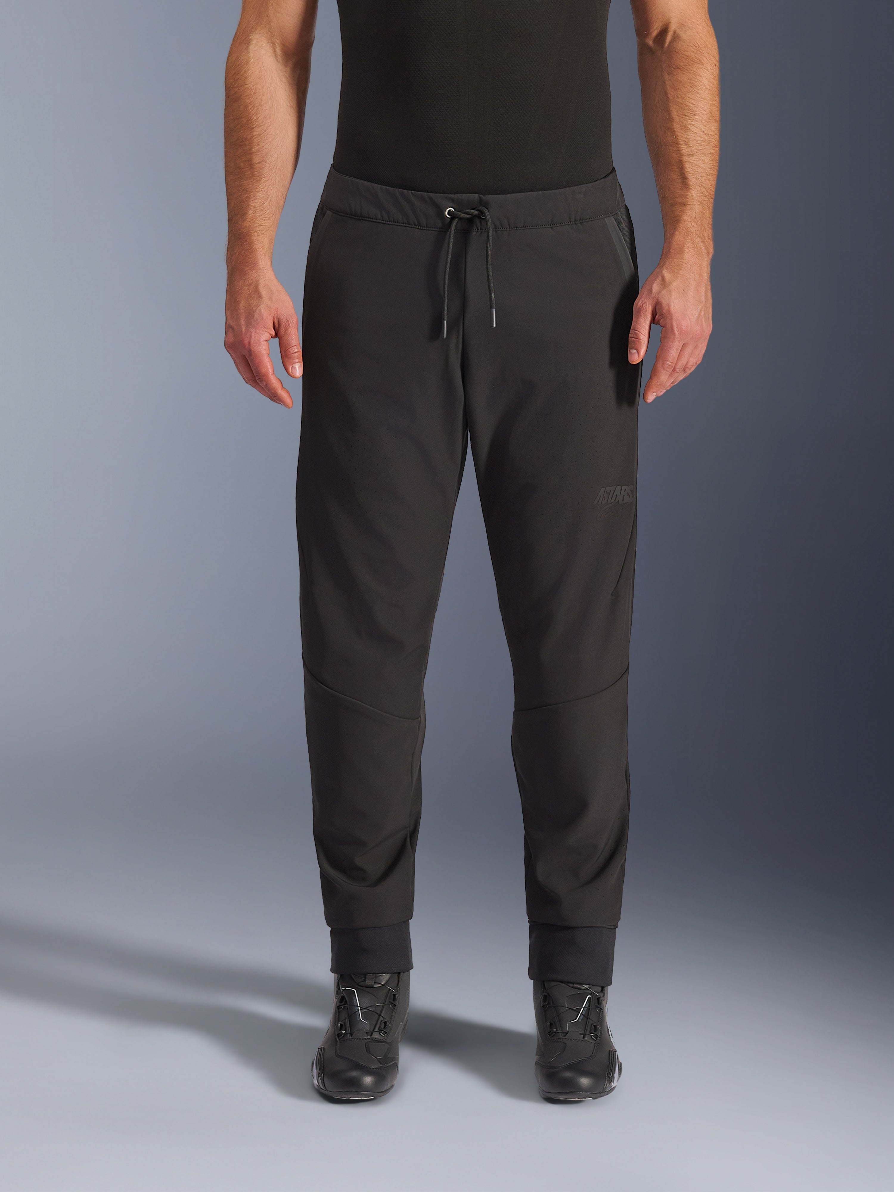 Pantaloni Alpinestars Aeroshell Airflow, pantaloni da moto neri, vista frontale indossati con stivali da moto neri, presentano vita con coulisse e polsini alle caviglie affusolati, design in softshell leggero con branding tonale discreto.