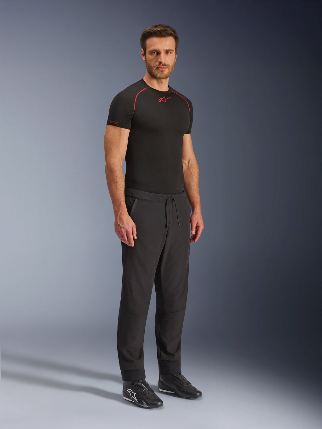 Alpinestars Aeroshell Airflow Pants, pantaloni da moto neri, scatto in studio a figura intera di un modello che indossa pantaloni in softshell con vita a coulisse e polsini elastici, abbinati a una maglia base layer Alpinestars nera e scarpe da guida nere.