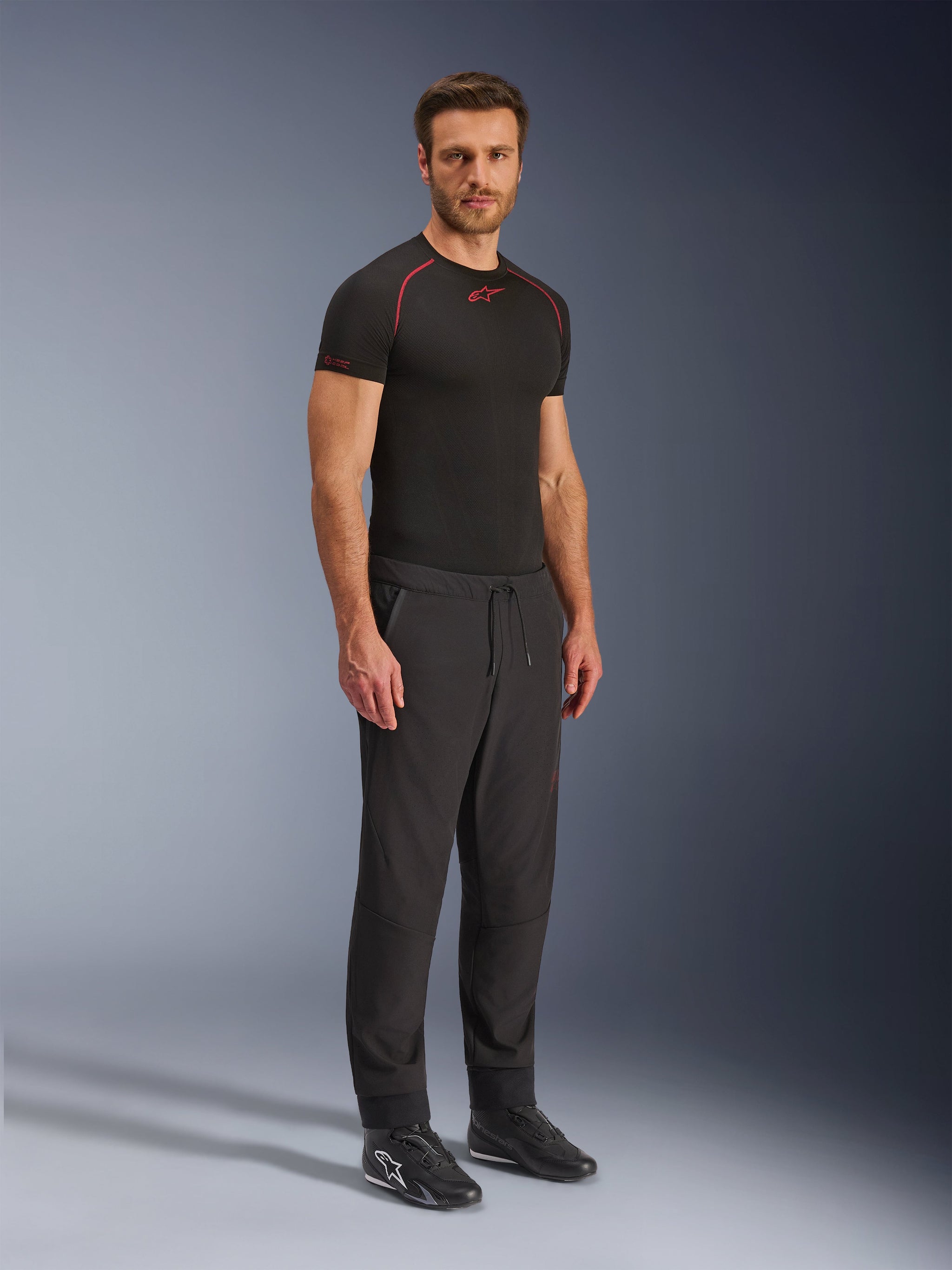 Alpinestars Aeroshell Airflow Pants, pantaloni da moto neri, scatto in studio a figura intera di un modello che indossa pantaloni in softshell con vita a coulisse e polsini elastici, abbinati a una maglia base layer Alpinestars nera e scarpe da guida nere.