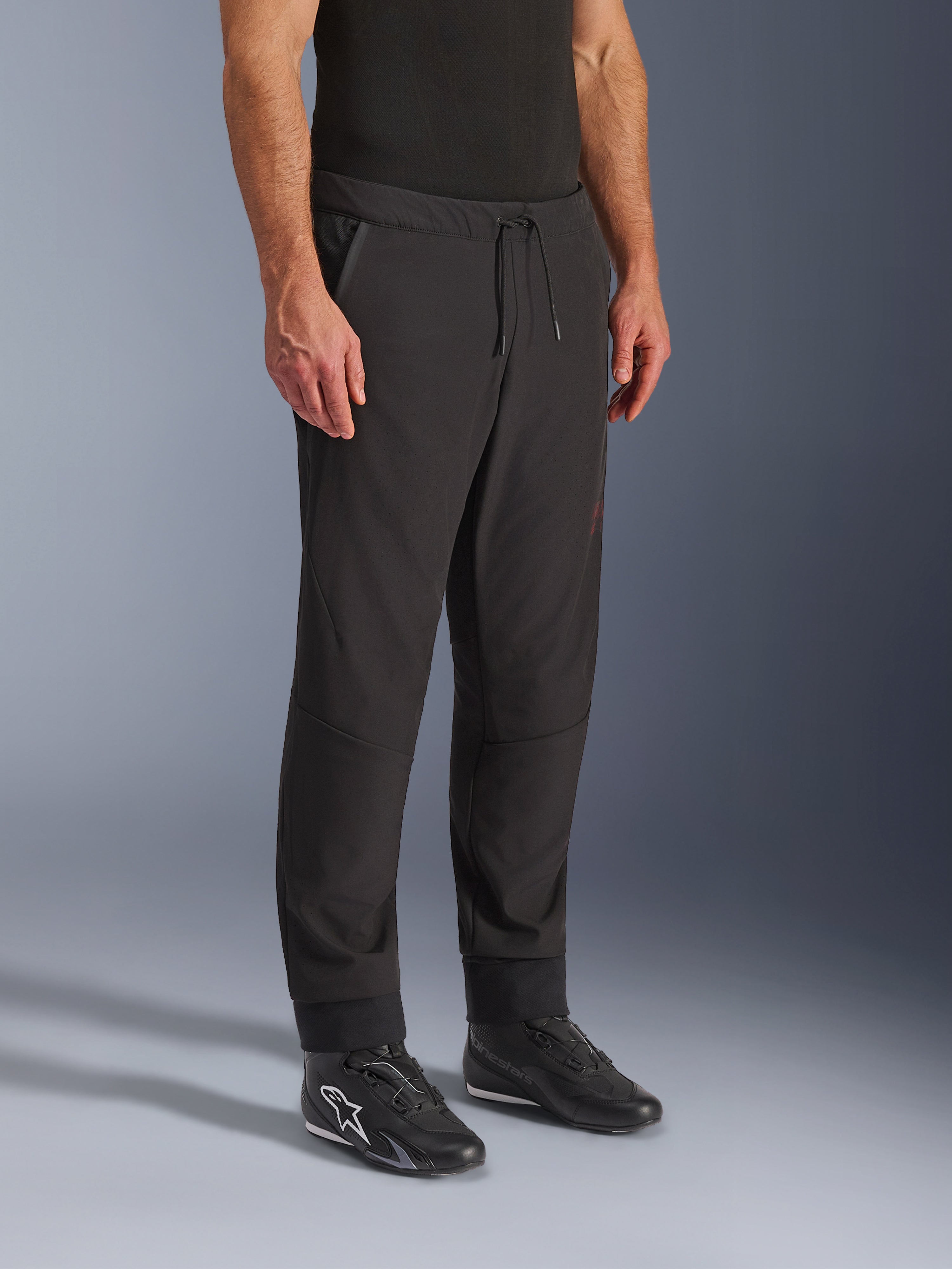 Pantaloni Alpinestars Aeroshell Airflow, pantaloni da moto neri, indossati con scarpe da guida nere, con vita a coulisse, polsini alle caviglie e tessuto softshell perforato al laser per la massima ventilazione
