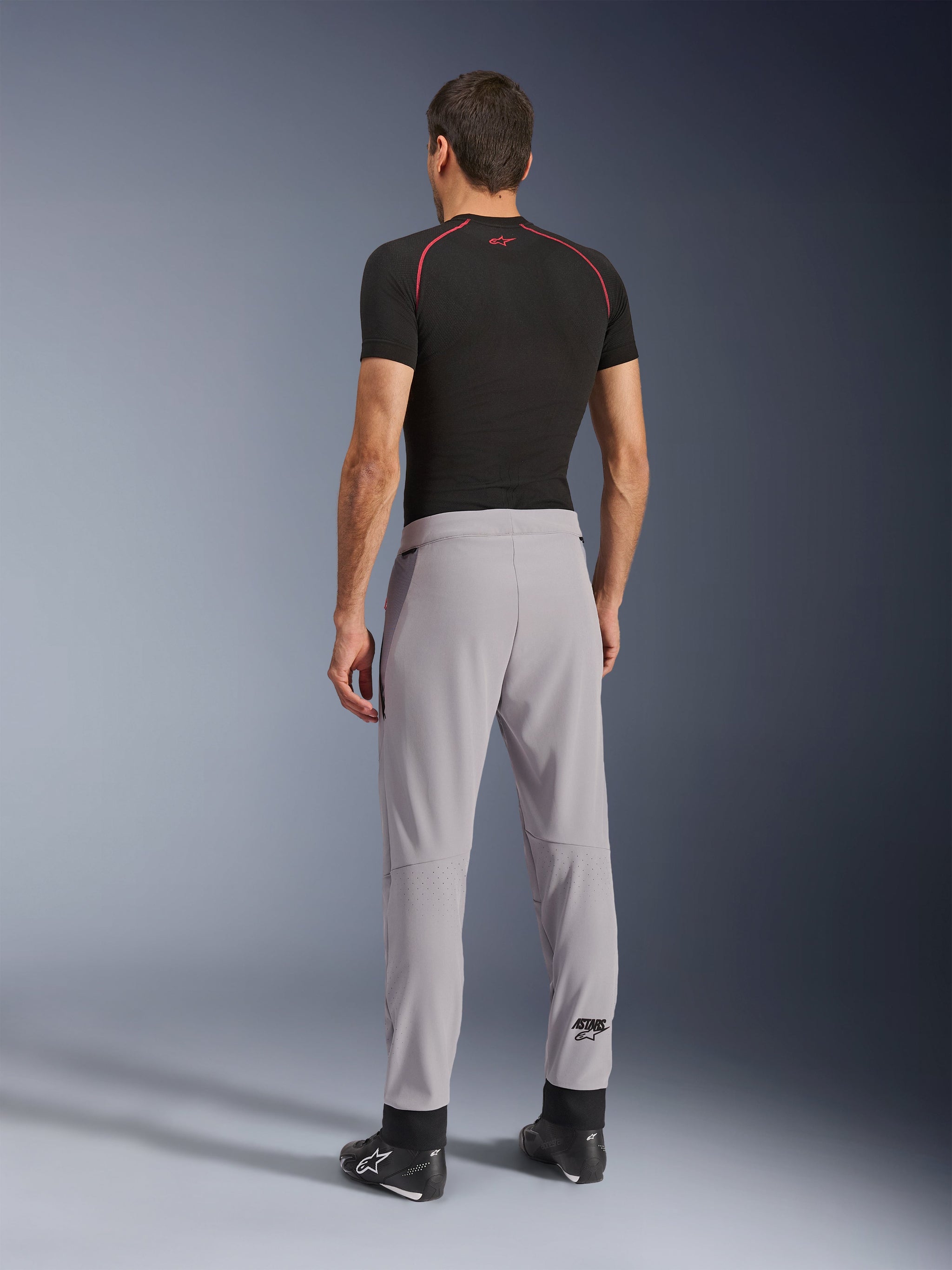 Vista posteriore di un modello che indossa i pantaloni Alpinestars Aeroshell Airflow in Mid Gray, abbinati a una maglia base layer nera e scarpe nere, che mette in risalto le zone laser-perforate sui polpacci e la vestibilità sportiva.