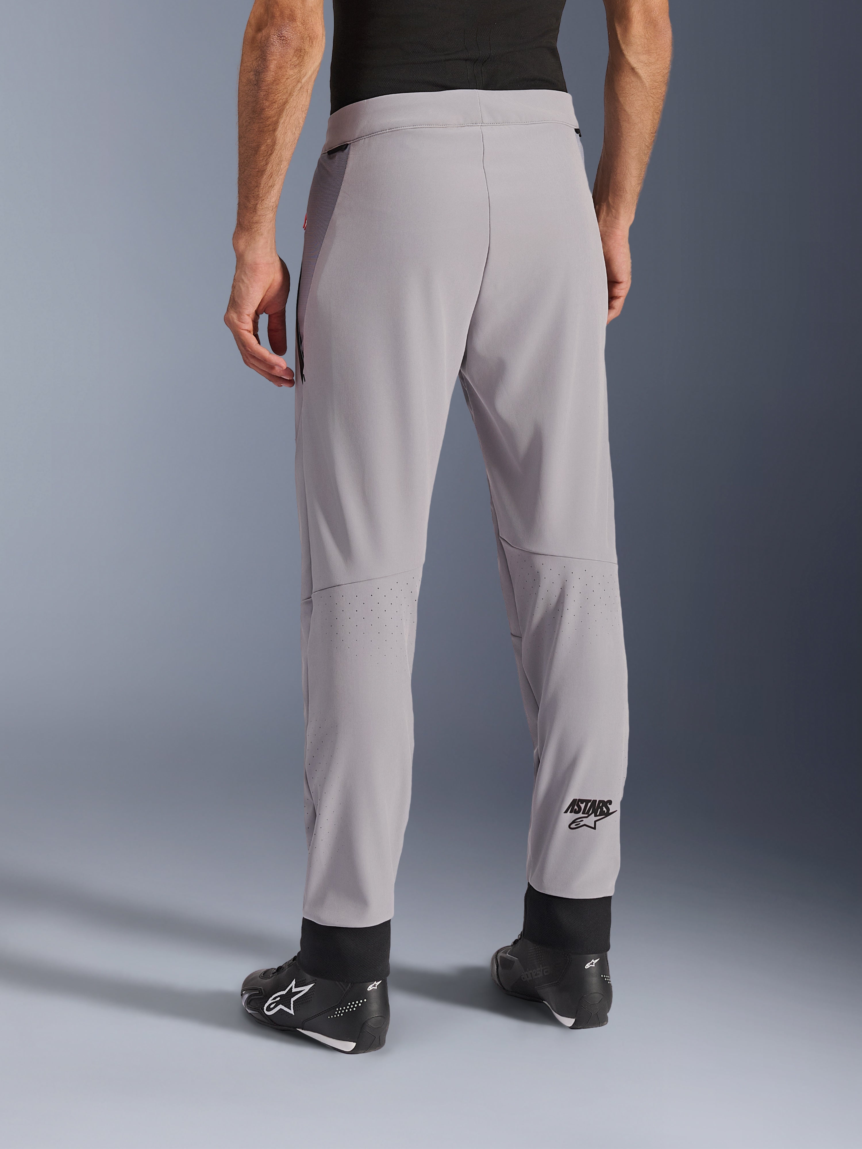 Pantaloni Alpinestars Aeroshell Airflow, Grigio Medio, vista posteriore che mostra le zone traforate al laser sui polpacci, logo ASTARS sulla parte inferiore della gamba, polsini neri regolabili alle caviglie, indossati con stivali da moto neri, pantaloni tecnici sportivi leggeri