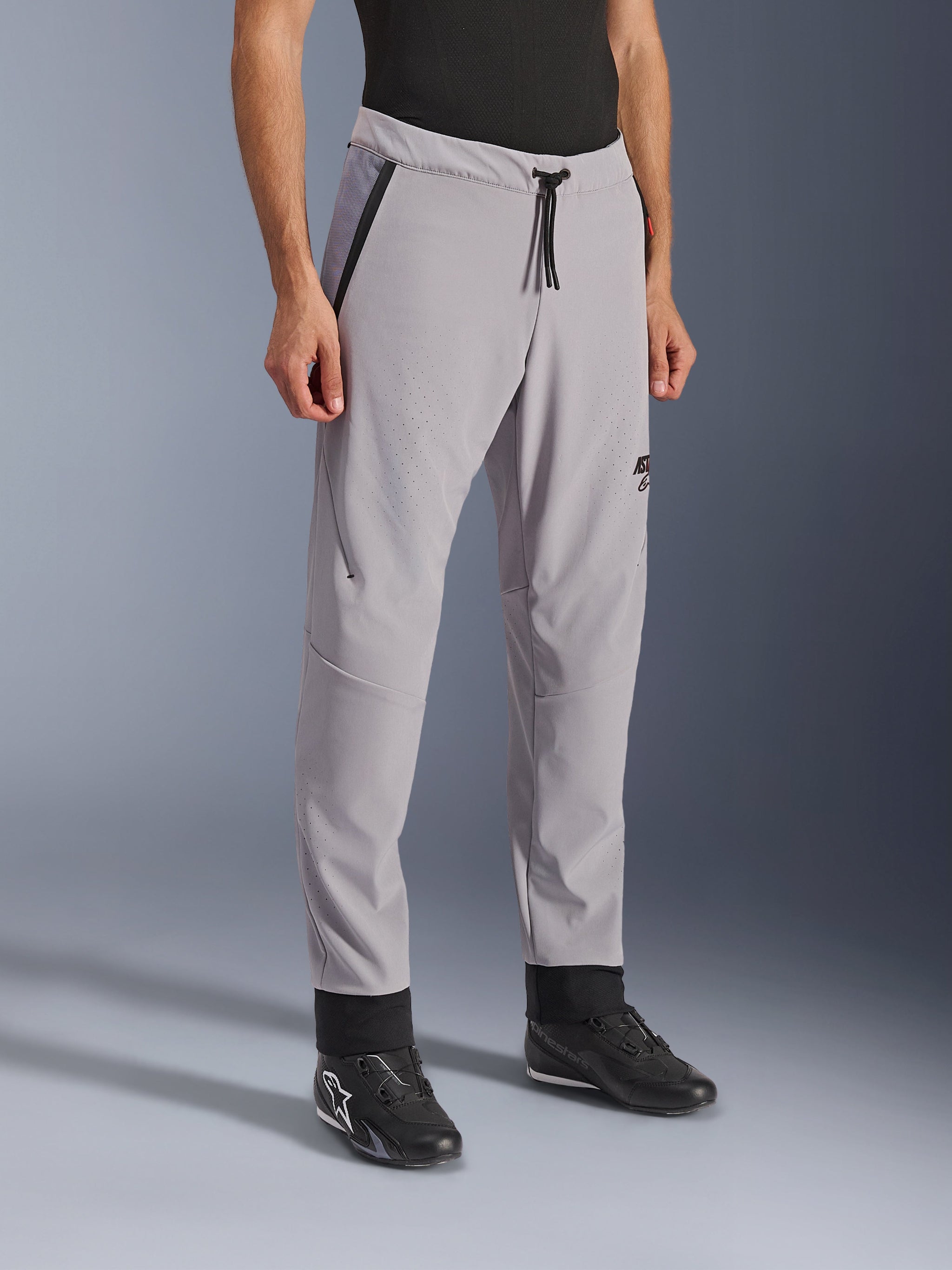 Pantaloni Alpinestars Aeroshell Airflow, grigio medio, pantaloni da moto leggeri con zone di ventilazione traforate al laser su cosce e stinchi, elastico in vita con cordino e vestibilità sportiva slim, indossati da un modello con scarpe da moto nere.
