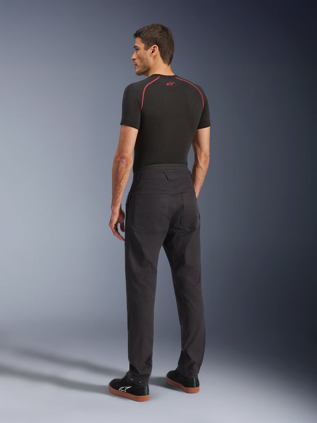 Alpinestars Flex-AST Canvas Pants, pantaloni da moto urbani neri, vista posteriore su modello, abbinati a un base layer nero a maniche corte e sneaker nere, caratterizzati da un design ispirato all'abbigliamento da lavoro con ampie tasche posteriori e un moderno taglio dritto.