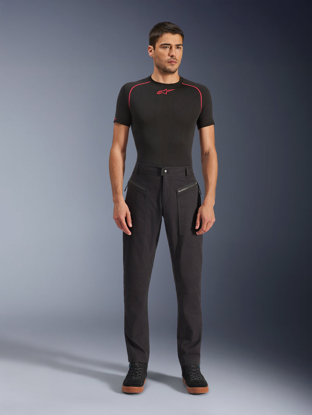 Pantaloni Alpinestars Flex-AST Canvas, pantaloni da moto urbani, nero, indossati da un modello con un base layer nero a maniche corte con cuciture rosse e sneakers casual nere, che mette in risalto le tasche anteriori con cerniera e il design ergonomico in tela.