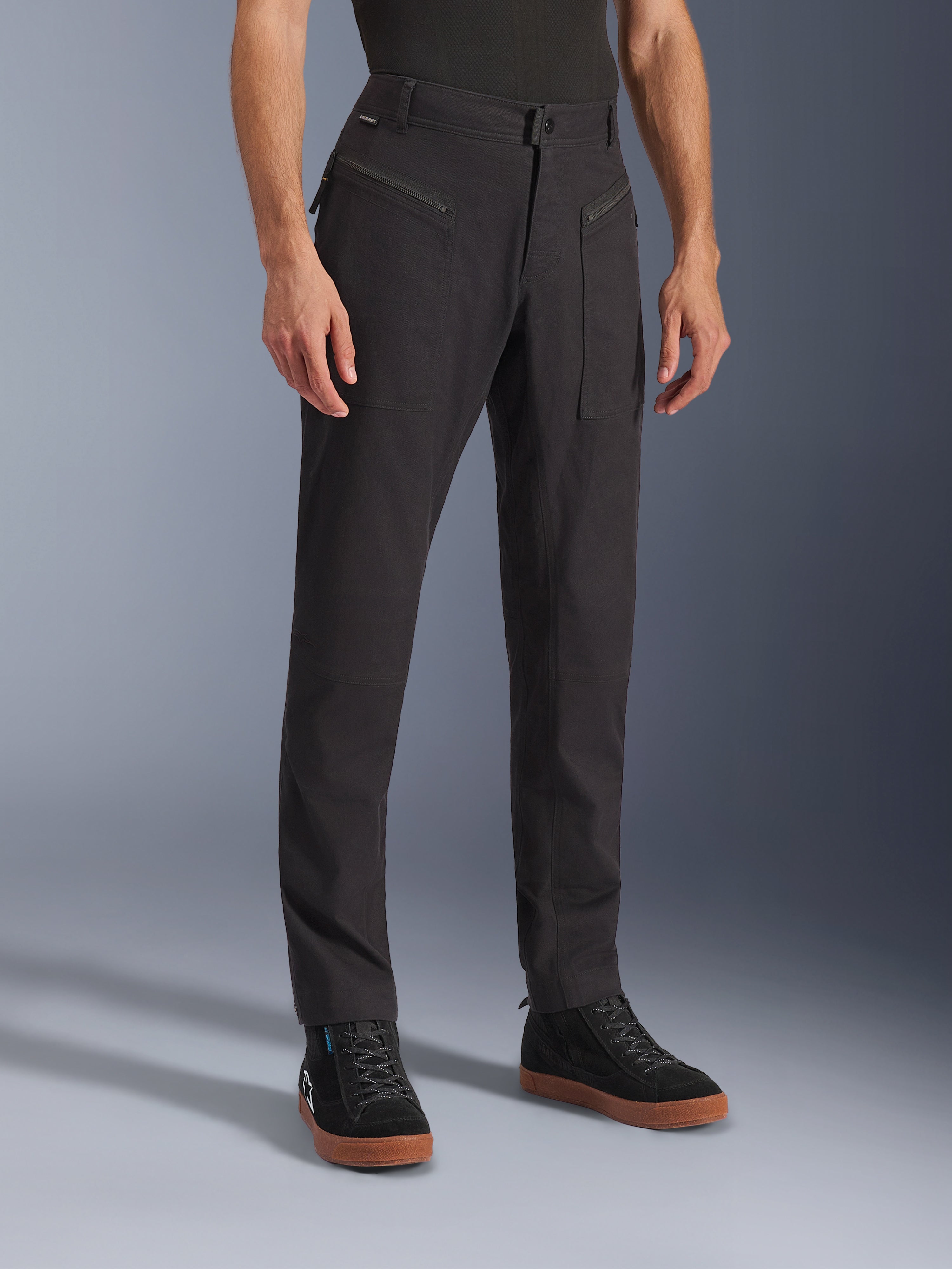 Alpinestars Flex-AST Canvas Pants, pantaloni da moto urbani, nero, indossati da un modello con sneaker nere, caratterizzati da un design workwear heritage, tessuto in twill di policotone elasticizzato e funzionali tasche anteriori con zip.