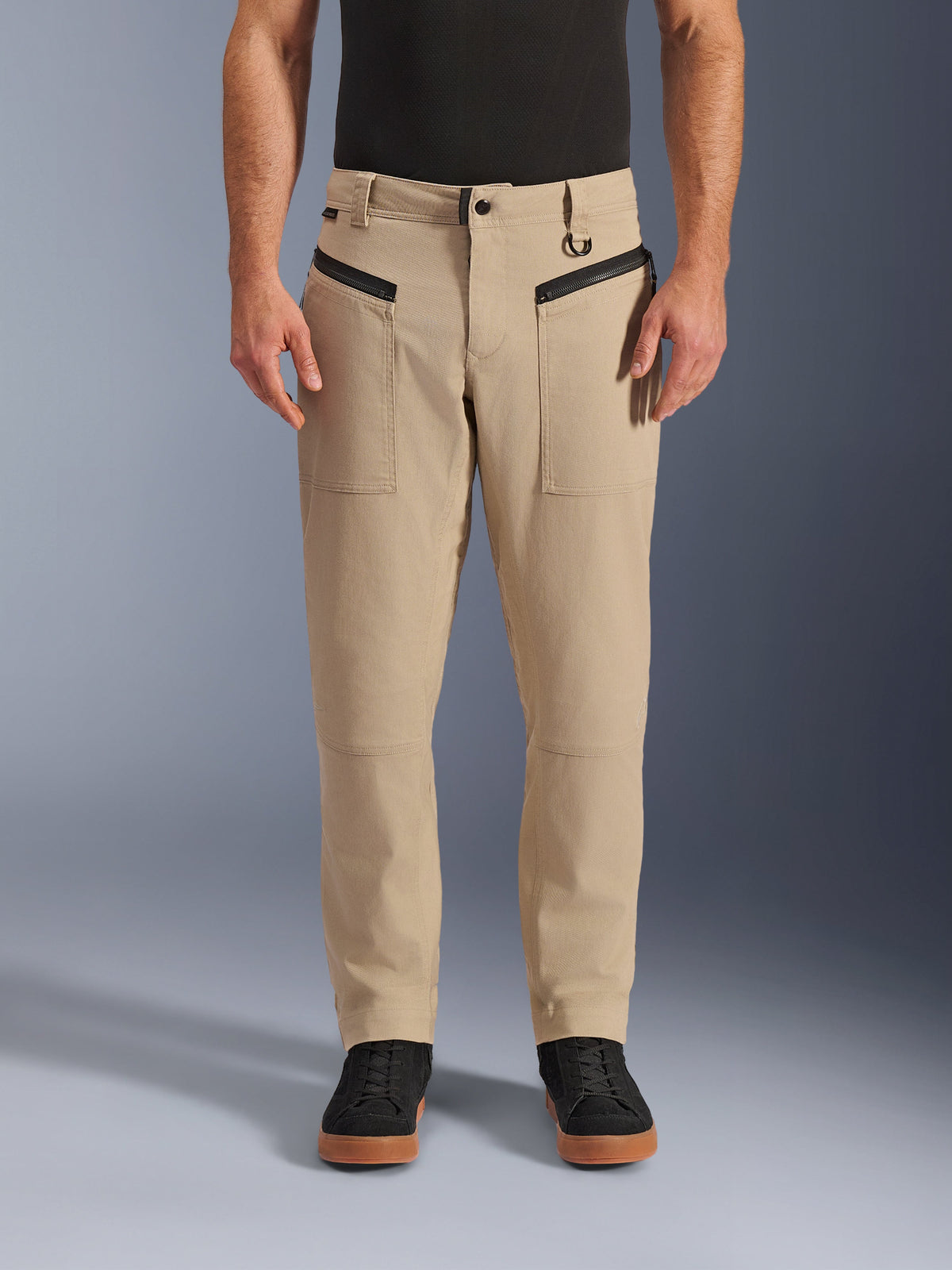 Alpinestars Pantaloni Flex-AST Canvas, pantaloni da moto urban, marrone, stile workwear heritage, con tasche anteriori orizzontali con zip nere e anello a D sul passante della cintura, indossati con maglia e scarpe nere, vista frontale in studio