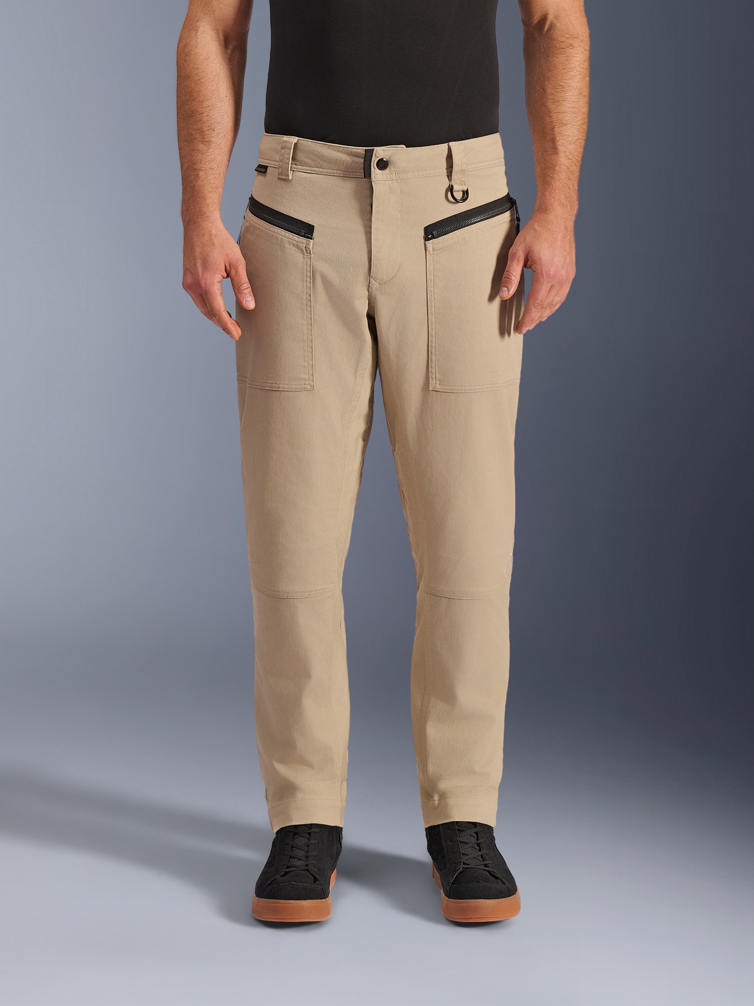 Alpinestars Pantaloni Flex-AST Canvas, pantaloni da moto urban, marrone, stile workwear heritage, con tasche anteriori orizzontali con zip nere e anello a D sul passante della cintura, indossati con maglia e scarpe nere, vista frontale in studio
