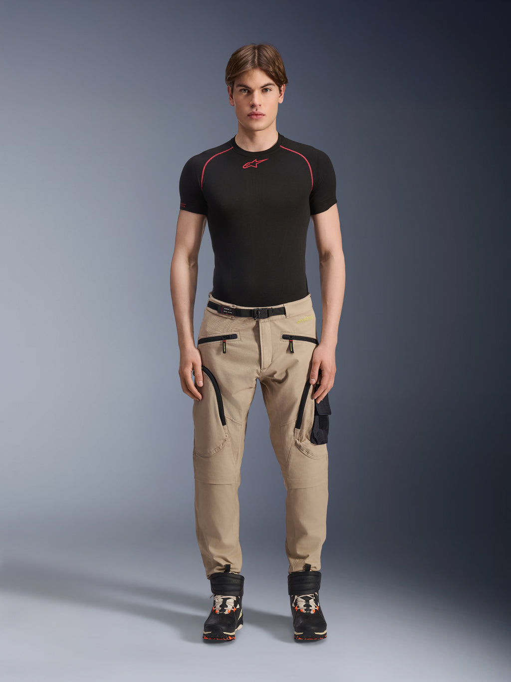 Modello che indossa pantaloni Alpinestars Acteon in colorazione Sand, abbinati a un base layer nero a maniche corte e stivali tecnici neri, con tasche cargo, zip nere e vestibilità affusolata, abbigliamento da moto urban contemporaneo