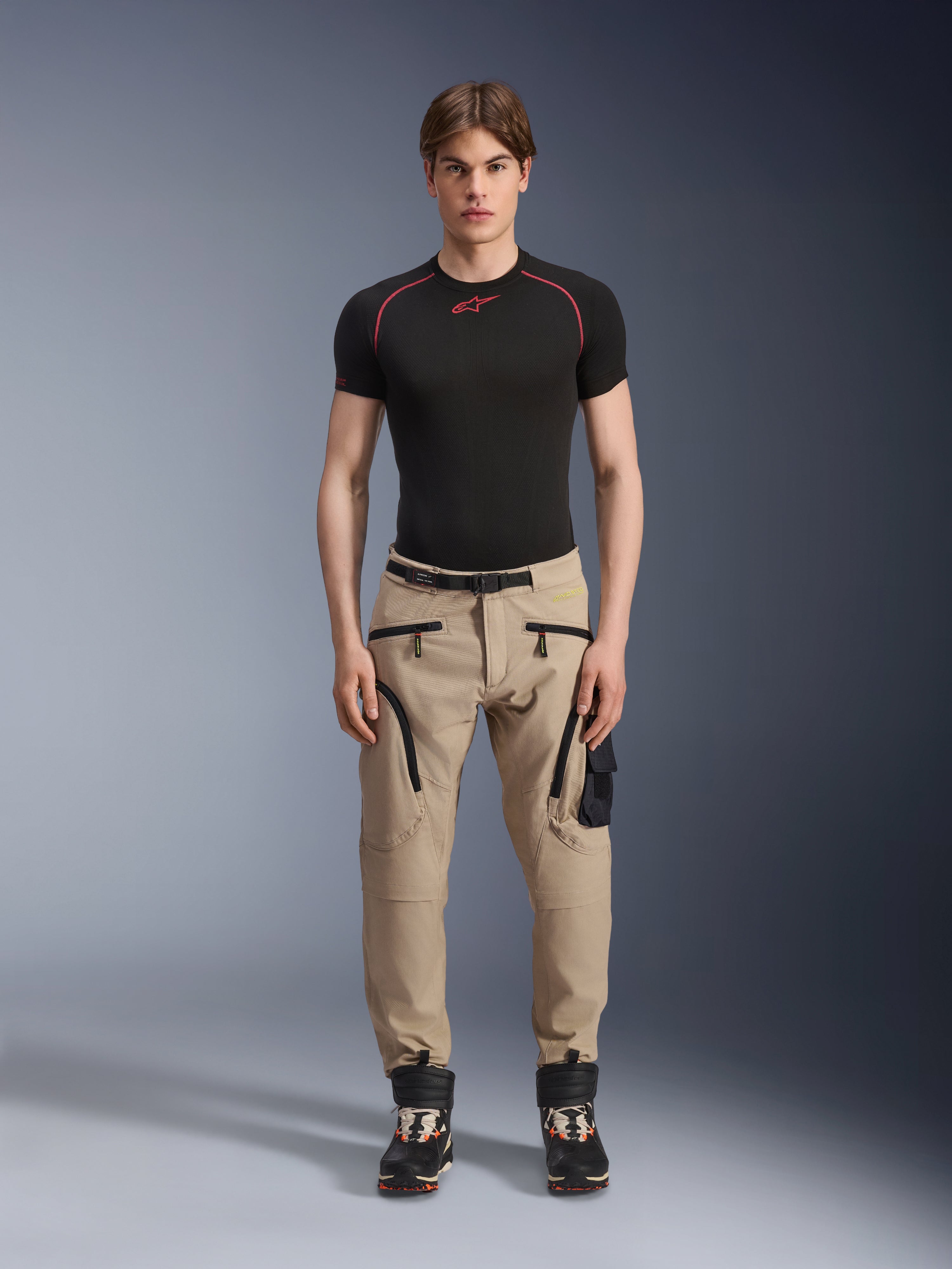 Modello che indossa pantaloni Alpinestars Acteon in colorazione Sand, abbinati a un base layer nero a maniche corte e stivali tecnici neri, con tasche cargo, zip nere e vestibilità affusolata, abbigliamento da moto urban contemporaneo